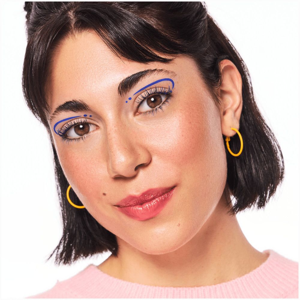 Femme avec un design d'eyeliner bleu autour des yeux. Boucles d'oreilles jaunes. Haut rose.