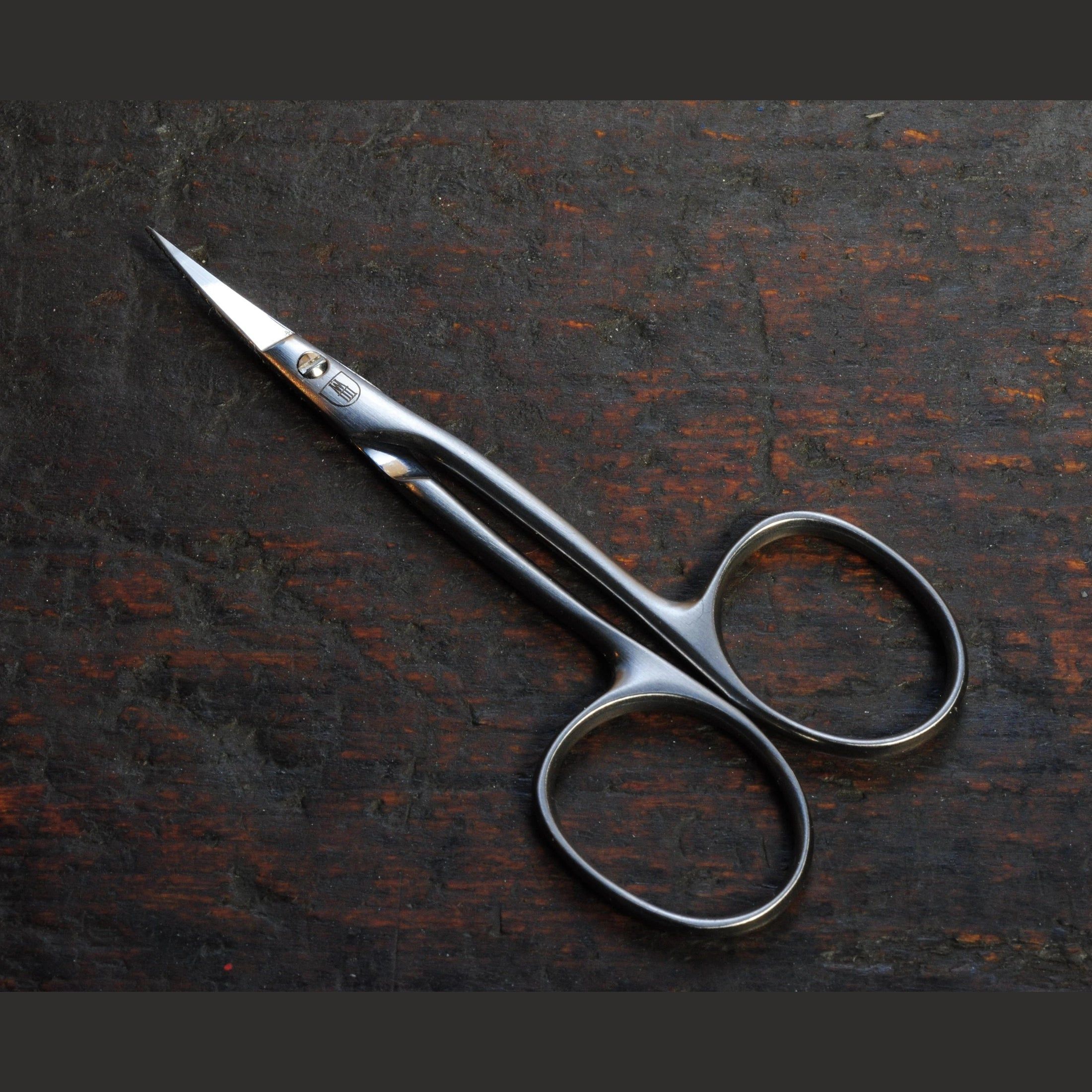 Ciseaux à cuticules en métal, sur bois sombre. Lames incurvées et poignées rondes.
