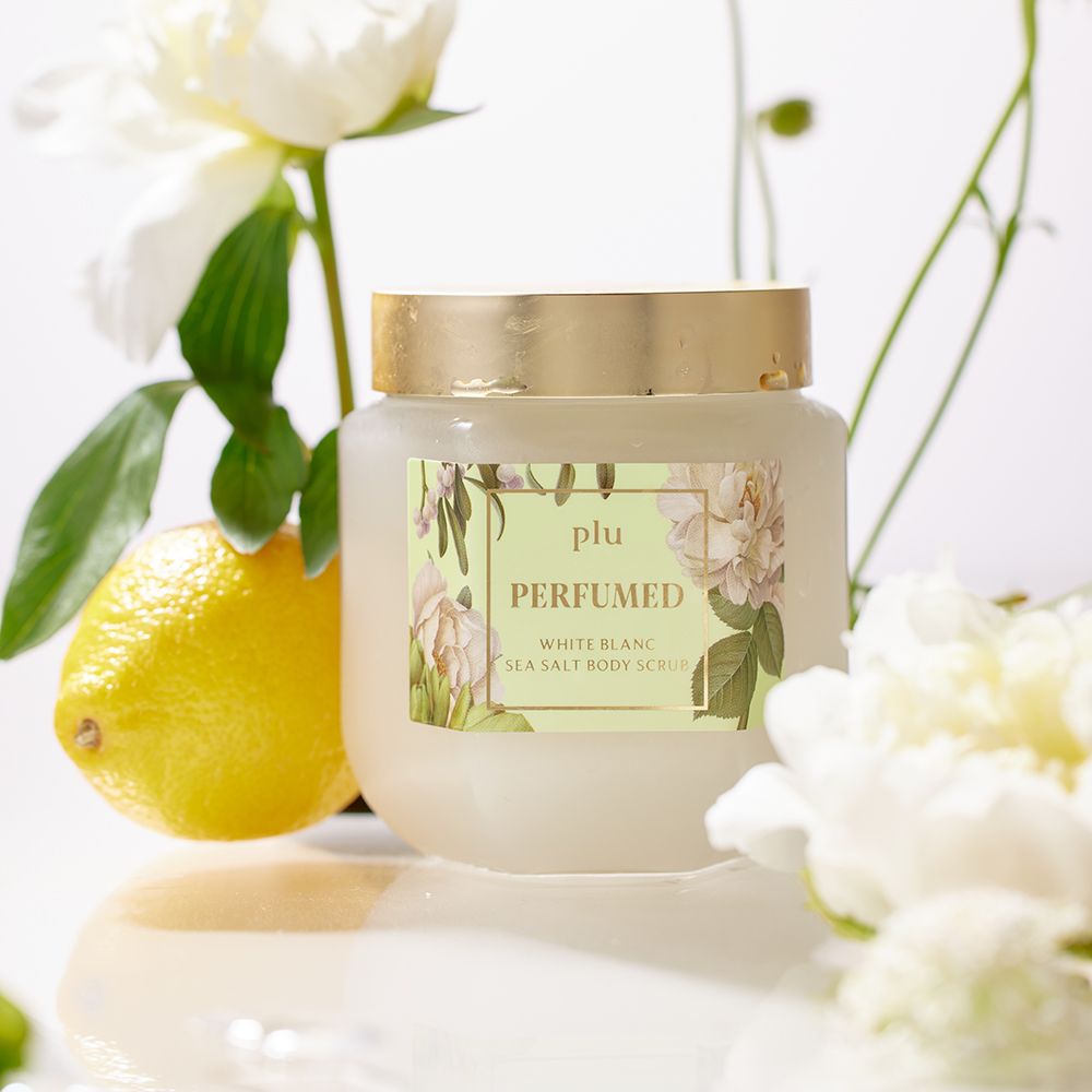Pot en verre avec couvercle doré, entouré de fleurs blanches et d'un citron. Étiquette avec nom du produit : PERFUMED White Blanc Sea Salt Body Scrub.