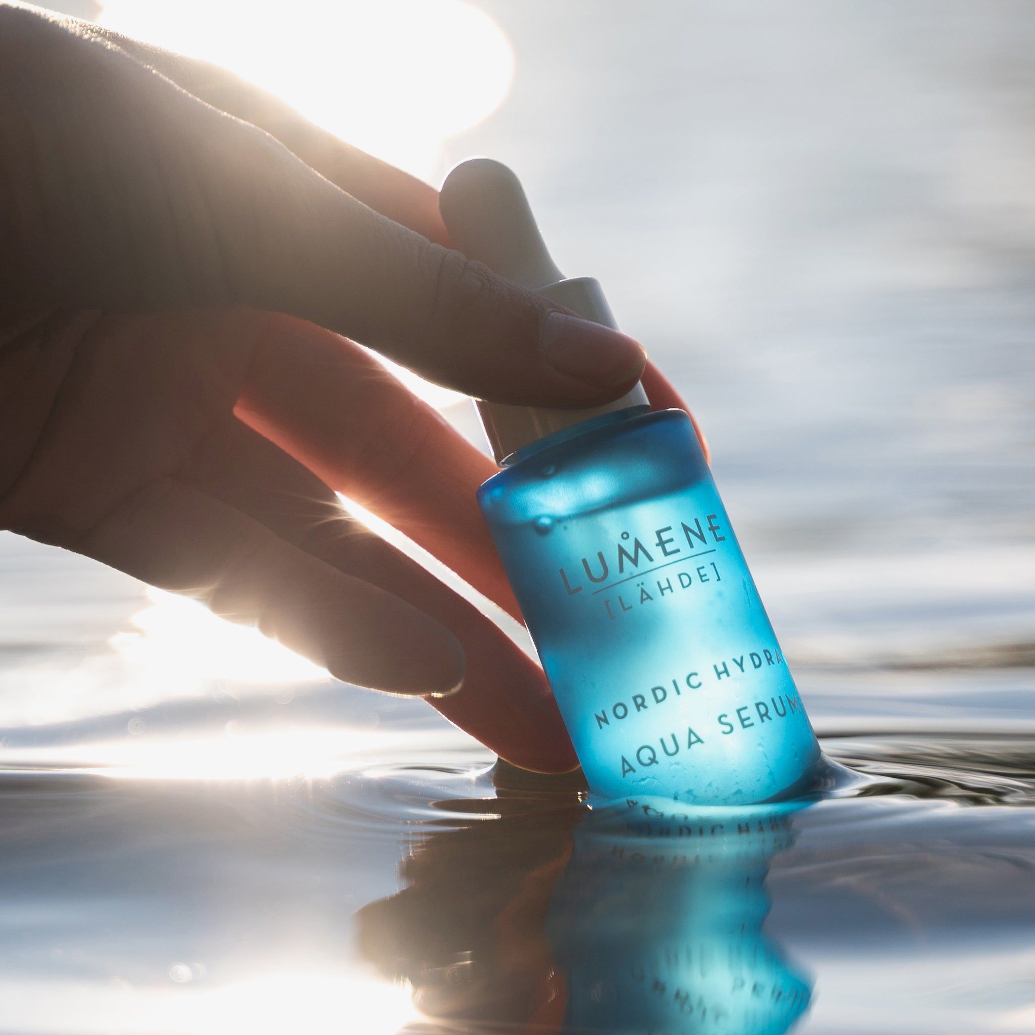 Flacon bleu dans l'eau, tenu par une main. Inscription : Lumene [LAHDE] Nordic Hydra Aqua Serum.