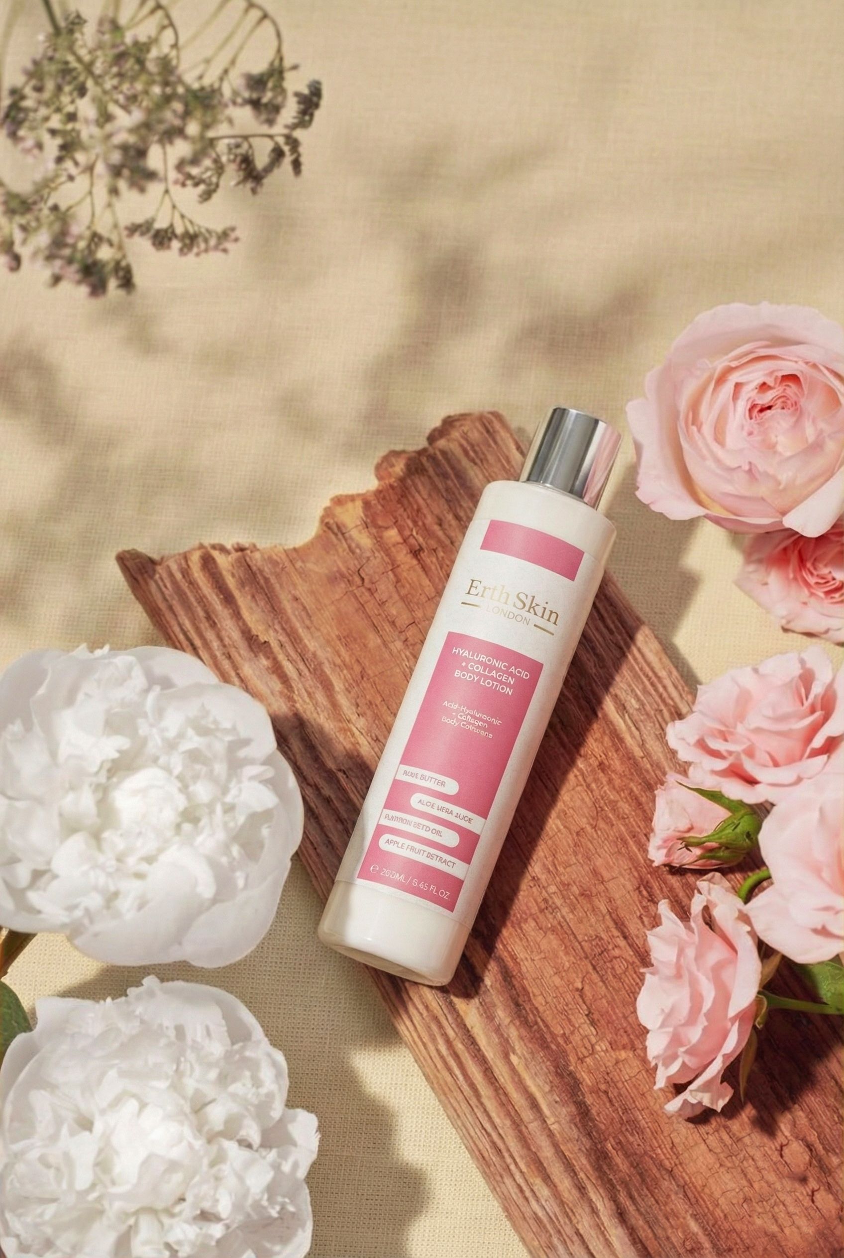 Flacon de lotion pour le corps blanc avec étiquette rose. Contient de l'acide hyaluronique et du collagène. Par ErthSkin London. Placé sur du bois et des fleurs.