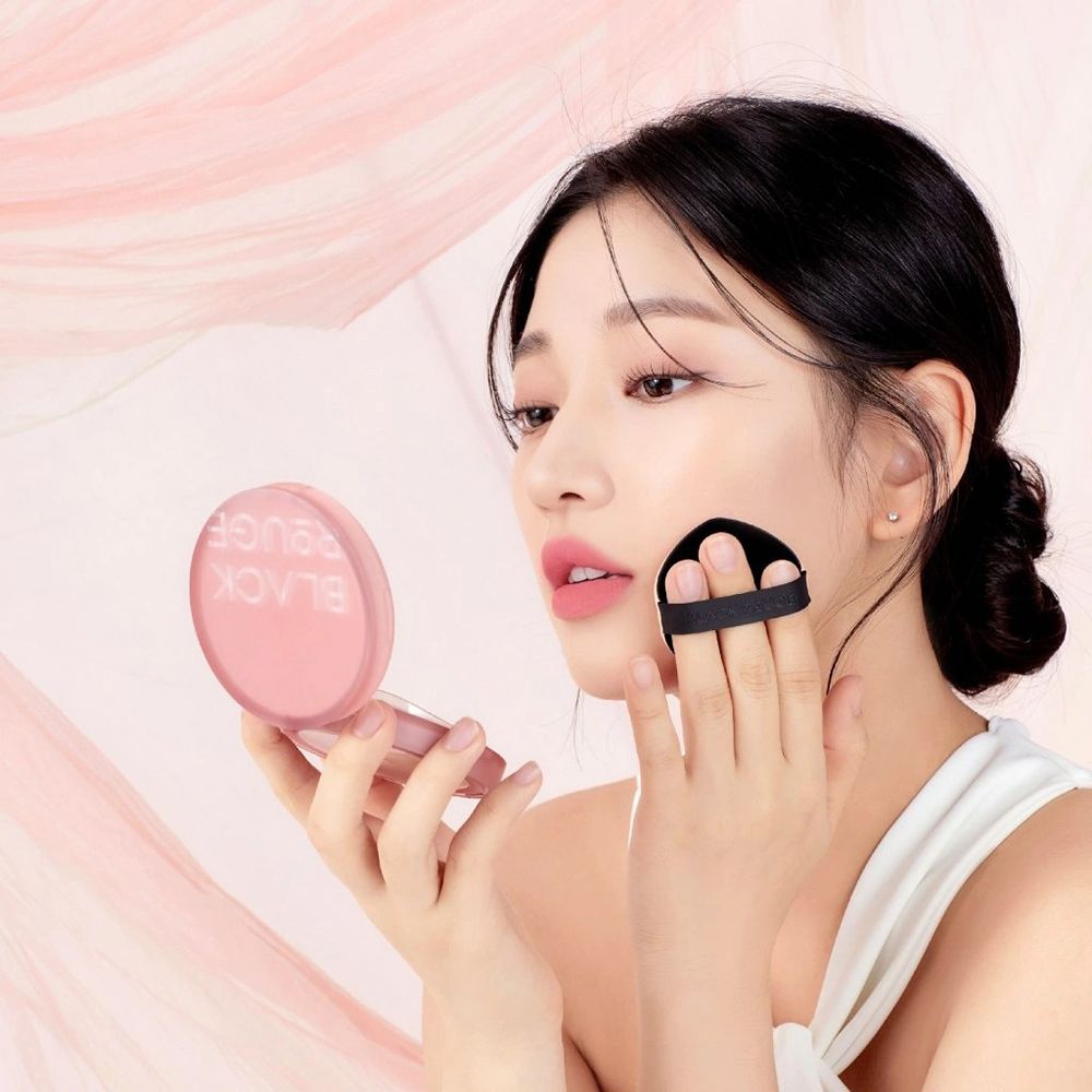 Femme appliquant le BLACK ROUGE Thin Layer Velour Cushion Foundation VC03 GINGER. Le produit et l'applicateur sont visibles.