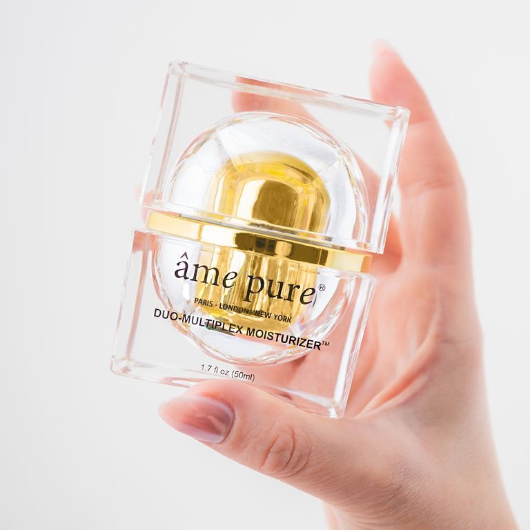 Een hand houdt een âme pure Duo-Multiplex Moisturizer vast in een transparante, vierkante verpakking met gouden details.
