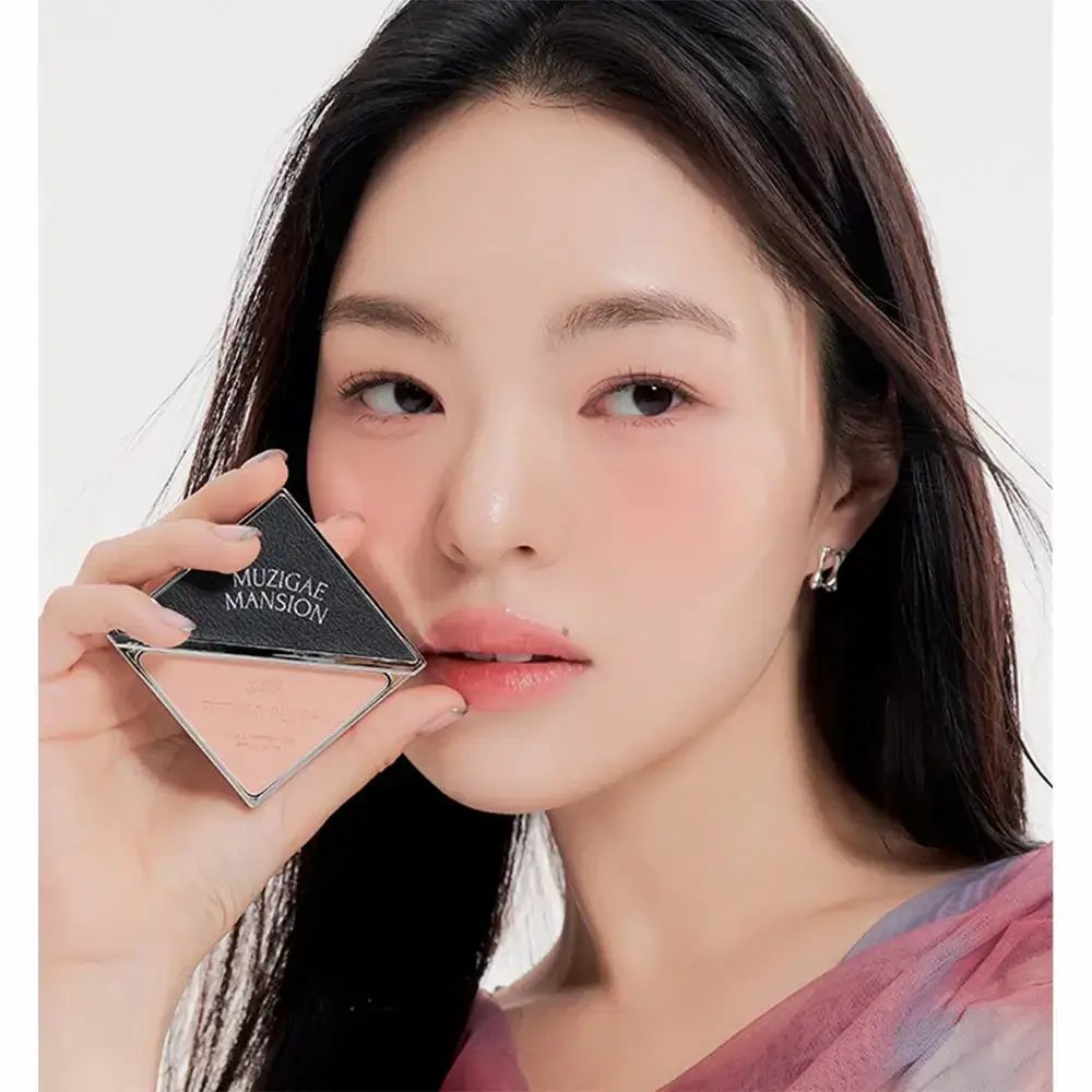 Vrouw houdt MUZIGAE MANSION blush vast. Vierkante, zilverkleurige compactverpakking. Roze blush zichtbaar. Vrouw draagt make-up.