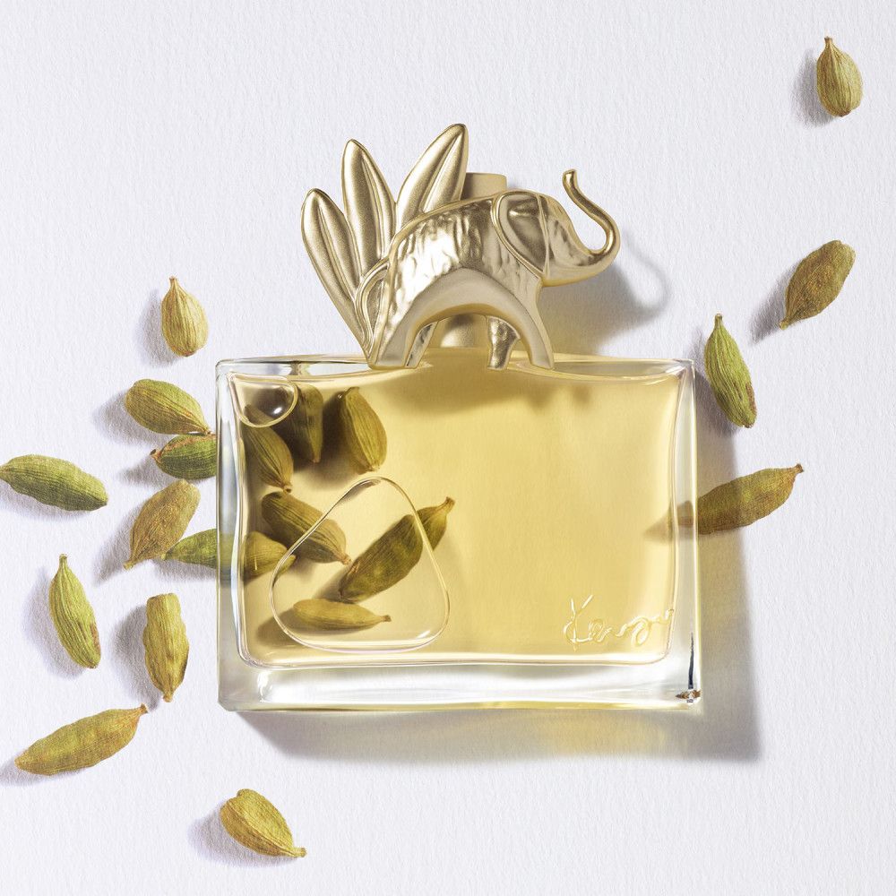 Flacon de parfum avec bouchon éléphant doré, entouré de gousses vertes. Récipient en verre clair avec liquide jaunâtre.