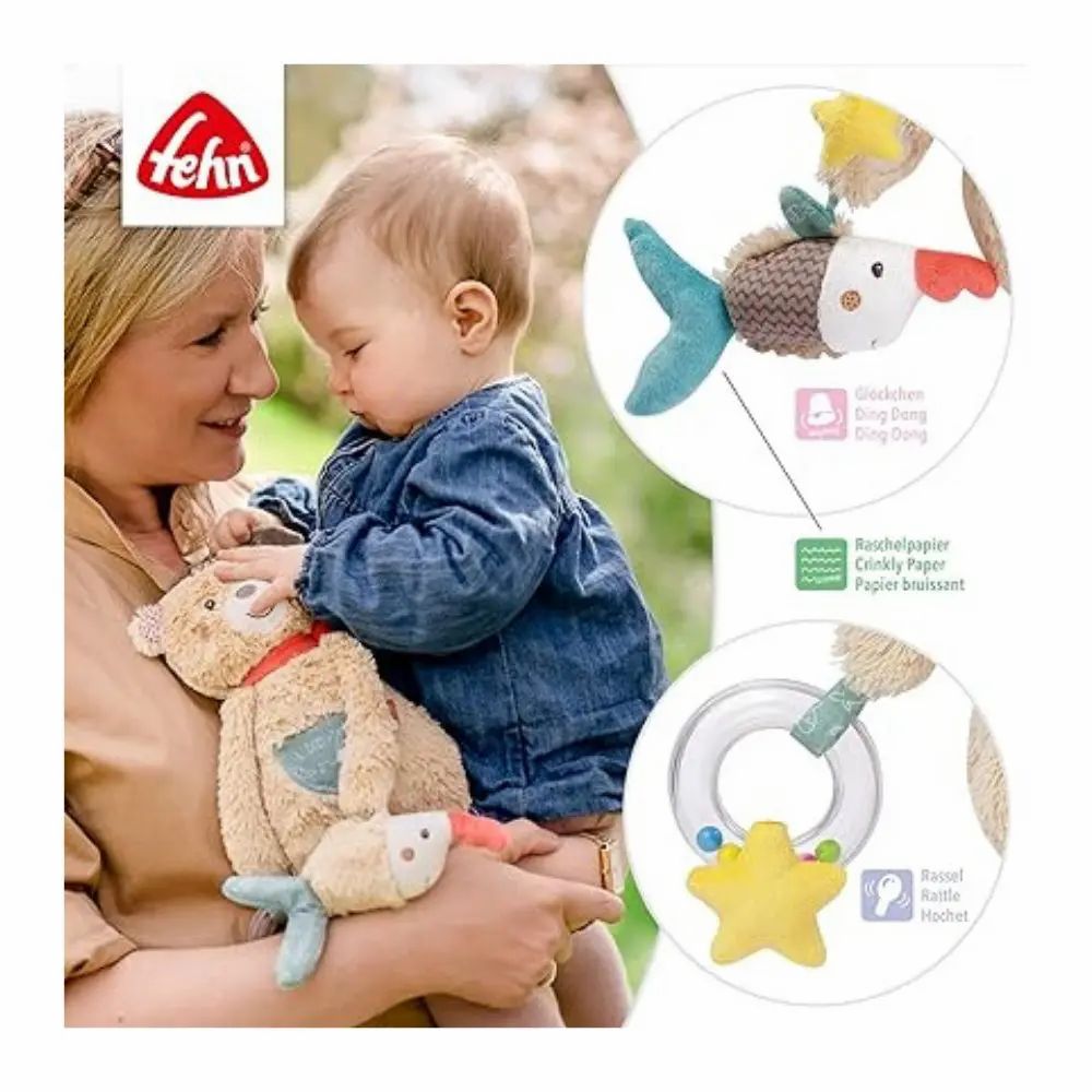Bébé tient peluche avec poisson. Peluche avec bonnet, écharpe rouge, étoile. Marque: fehn. Avec texte.
