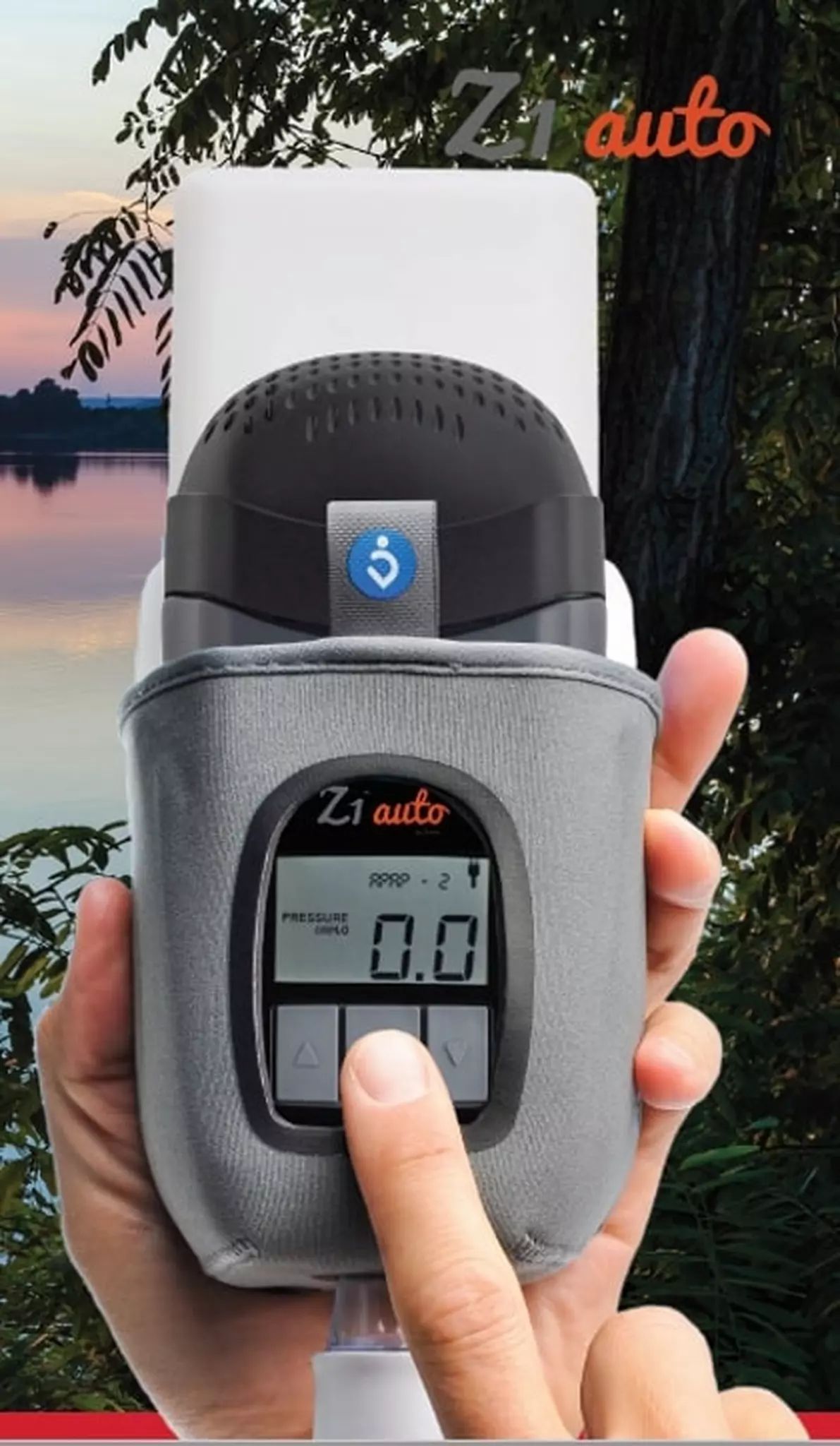 Appareil CPAP de voyage gris avec écran, tenu par des mains, devant un lac et des arbres.