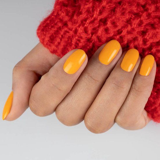 Main avec ongles vernis en orange. Les ongles sont de forme ovale. Pull rouge en arrière-plan.