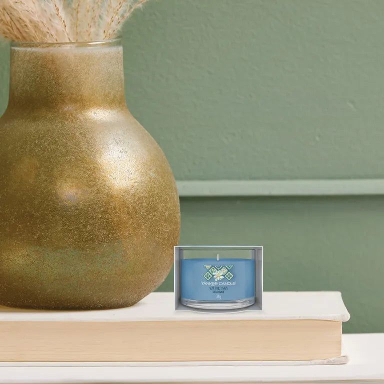 Bougie bleu clair dans verre, sur livre et à côté vase. Inscription: Yankee Candle Azure Sky, 37g.