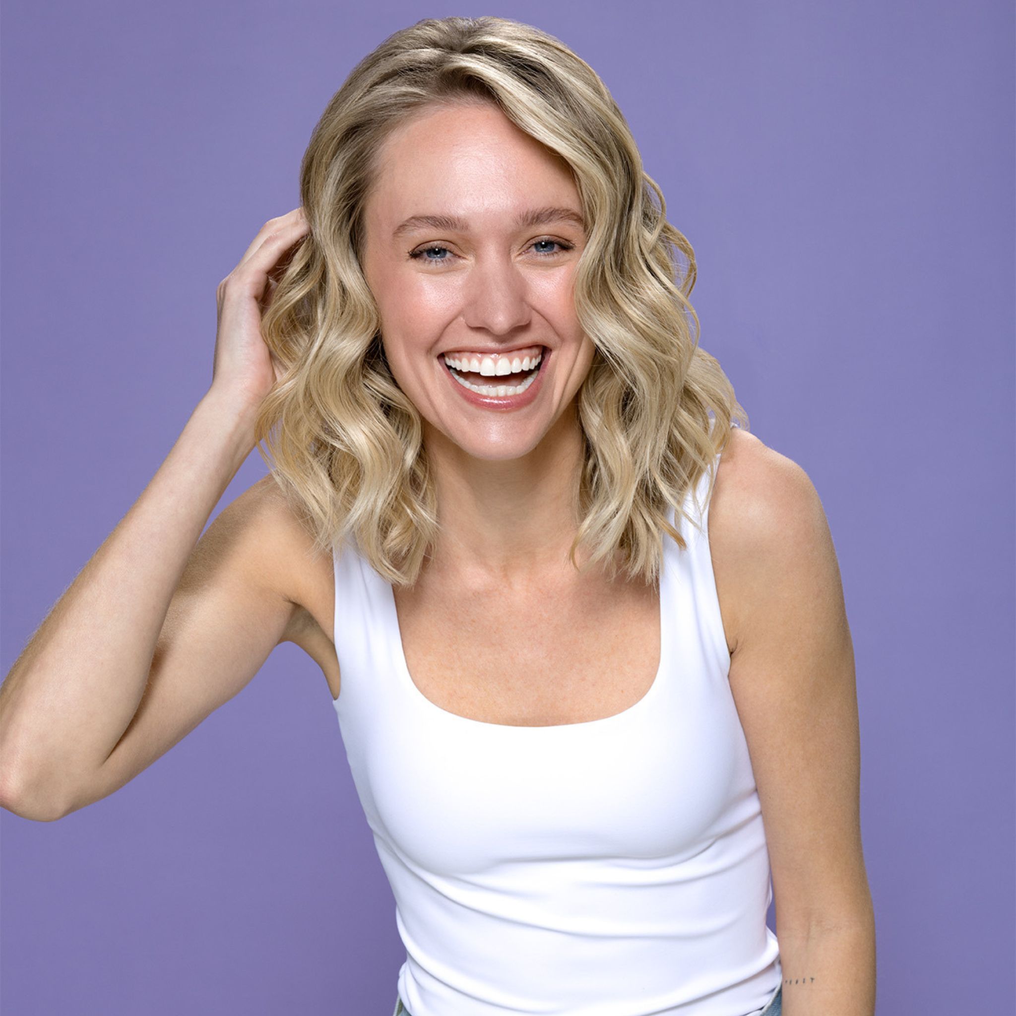 Lachende vrouw met blond haar. Ze draagt een witte tanktop. Paarse achtergrond.
