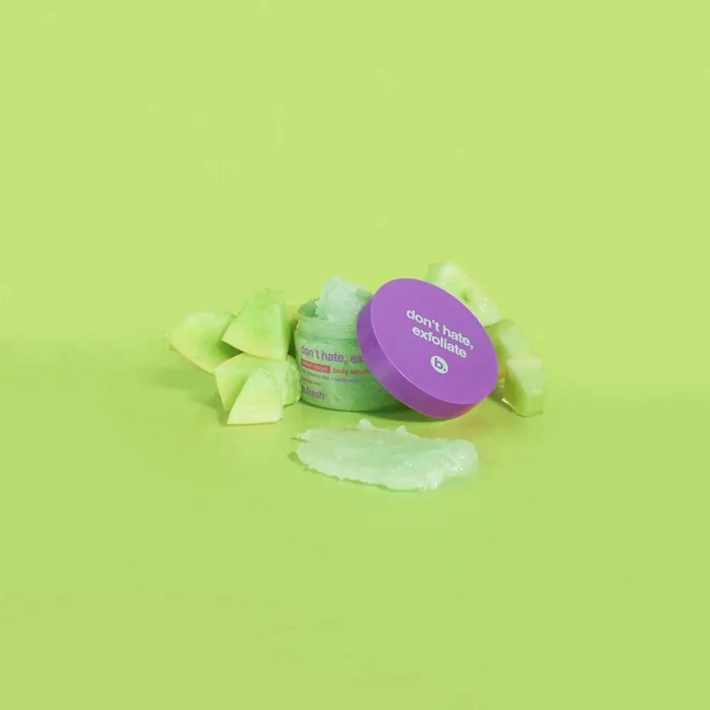 Open lichaamsscrubpot met paarse deksel en meloenschijfjes op groene achtergrond. Productnaam zichtbaar op de deksel.