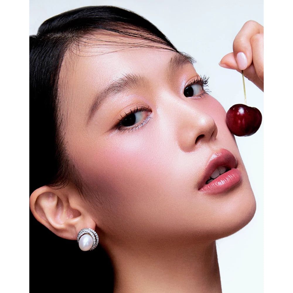 Portrait d'une femme avec du fard à joues. Elle tient une cerise. Elle porte des boucles d'oreilles.