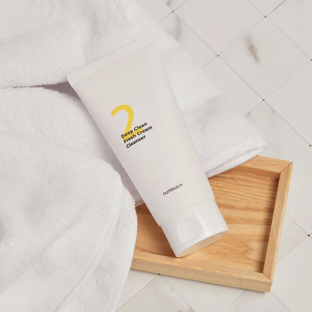 Witte tube met gele "2" en tekst "Deep Clean Fresh Cream Cleanser" op houten dienblad en handdoek.