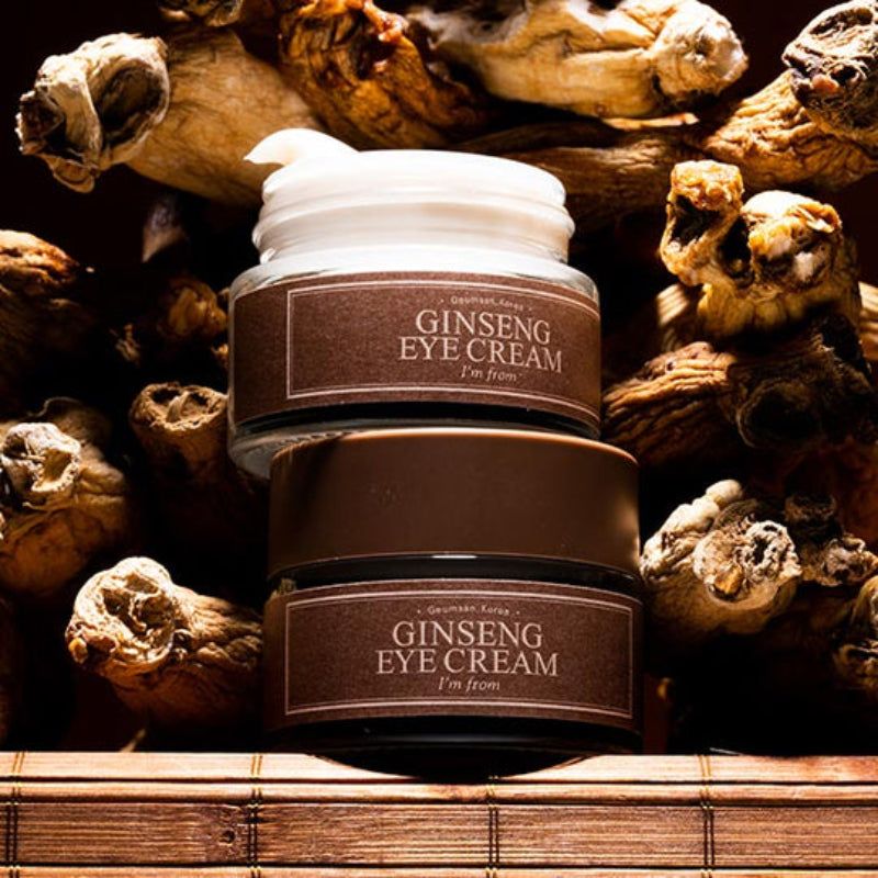 Twee potjes oogcrème, bruine deksels, etiketten. Tekst: Ginseng Eye Cream, I'm from. Achtergrond: wortels.