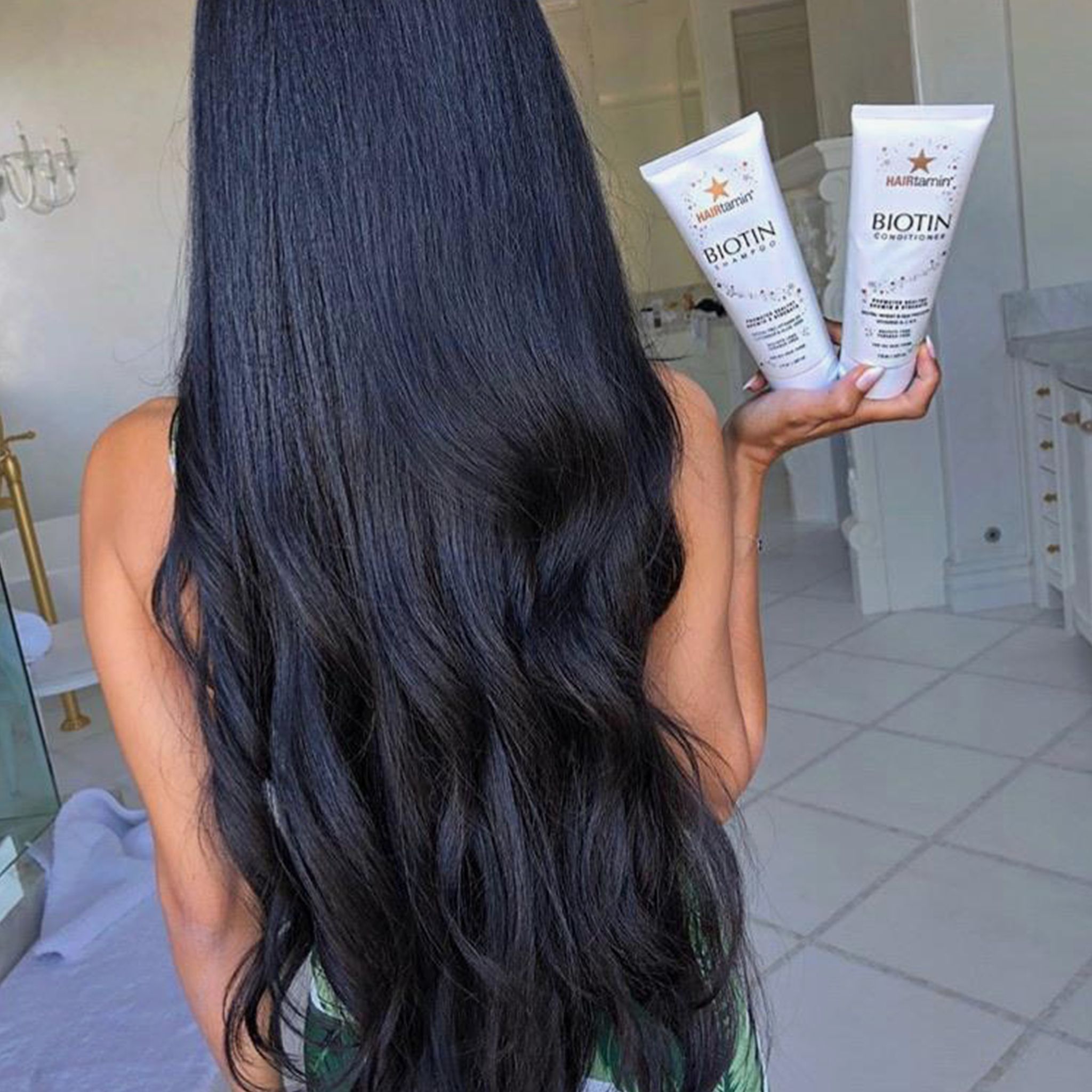 Personne tenant deux tubes "HAIRtamin Biotin Conditioner". Longs cheveux noirs au premier plan. Arrière-plan salle de bain.