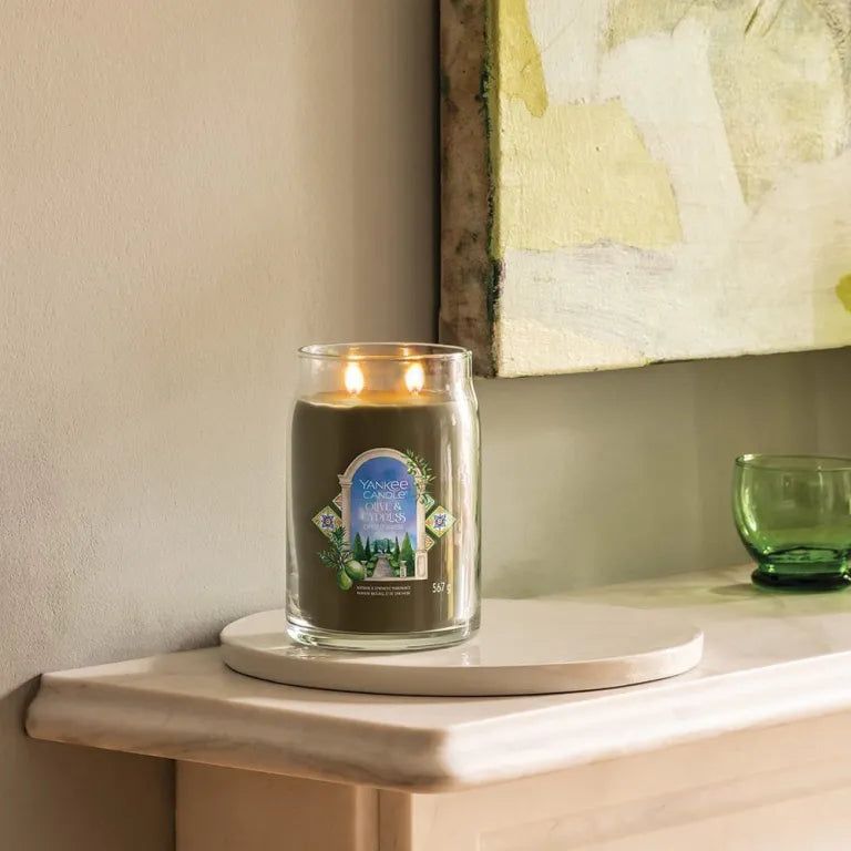 Brandende Yankee Candle Olive & Cypress in glazen pot. Op wit dienblad, voor muur met kunstwerk.