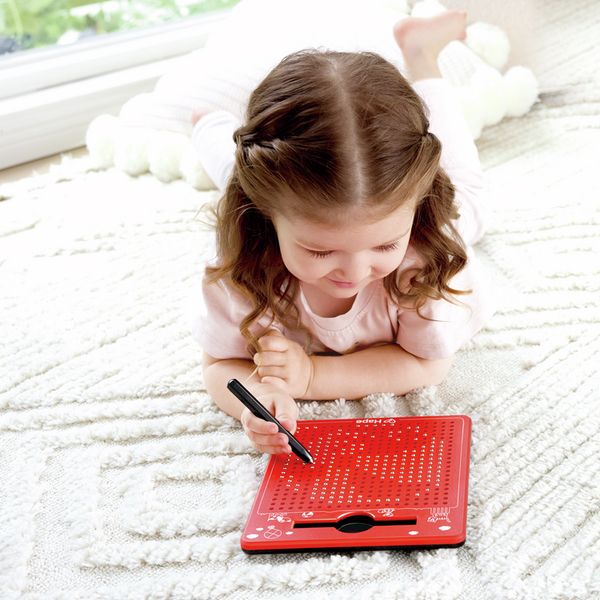 Klein meisje tekent met een pen op een rode Hape magnetische tekentafel. Kind ligt op een tapijt.