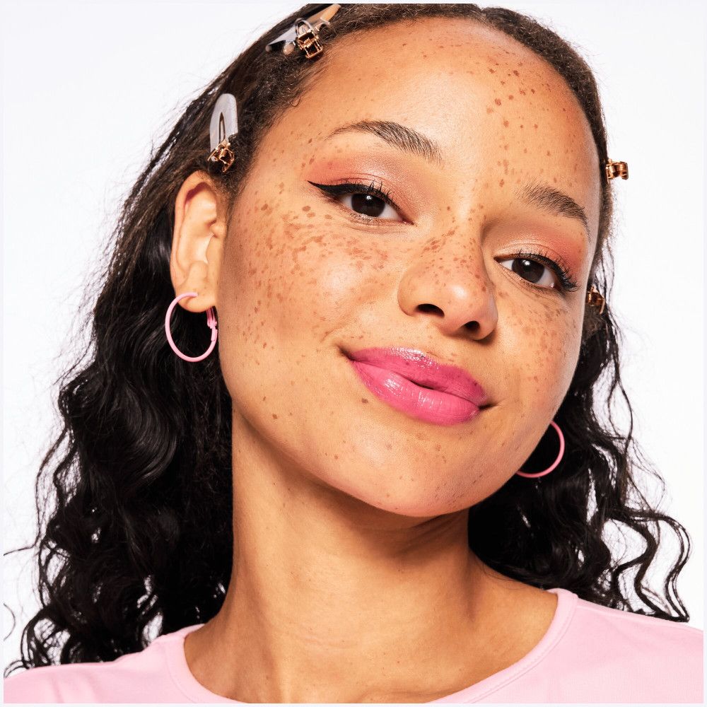 Portrait d'une femme avec du baume à lèvres rose. Elle porte des créoles roses et un haut rose.