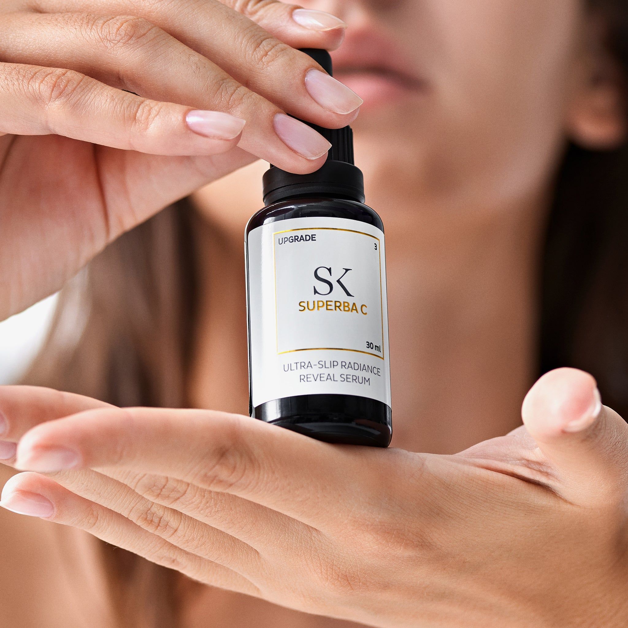 Handen houden een zwarte fles met pipet. Op het etiket staat "SK SUPERBA C" en "ULTRA-SLIP RADIANCE REVEAL SERUM".
