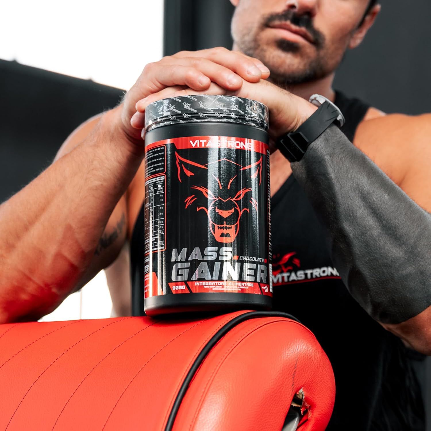 Conteneur Vitastrong Mass Gainer. Conteneur noir avec étiquette rouge. Le conteneur est tenu par une personne.