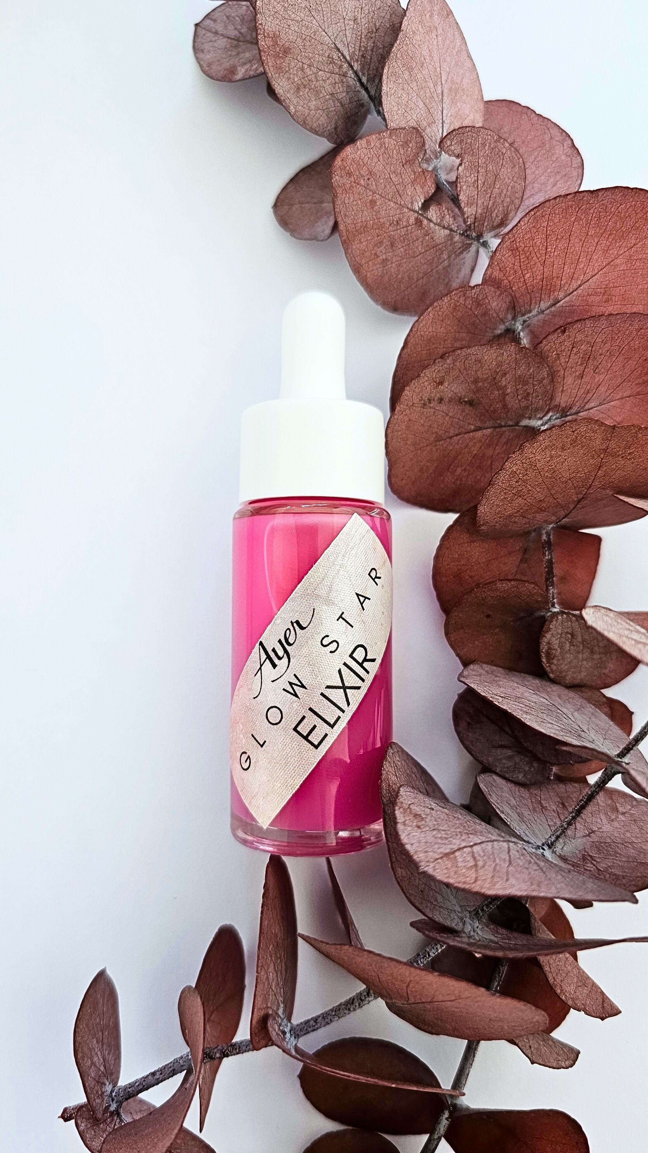 Flacon de liquide rose avec compte-gouttes blanc, entouré de feuilles séchées. Étiquette: Glow Star Elixir.