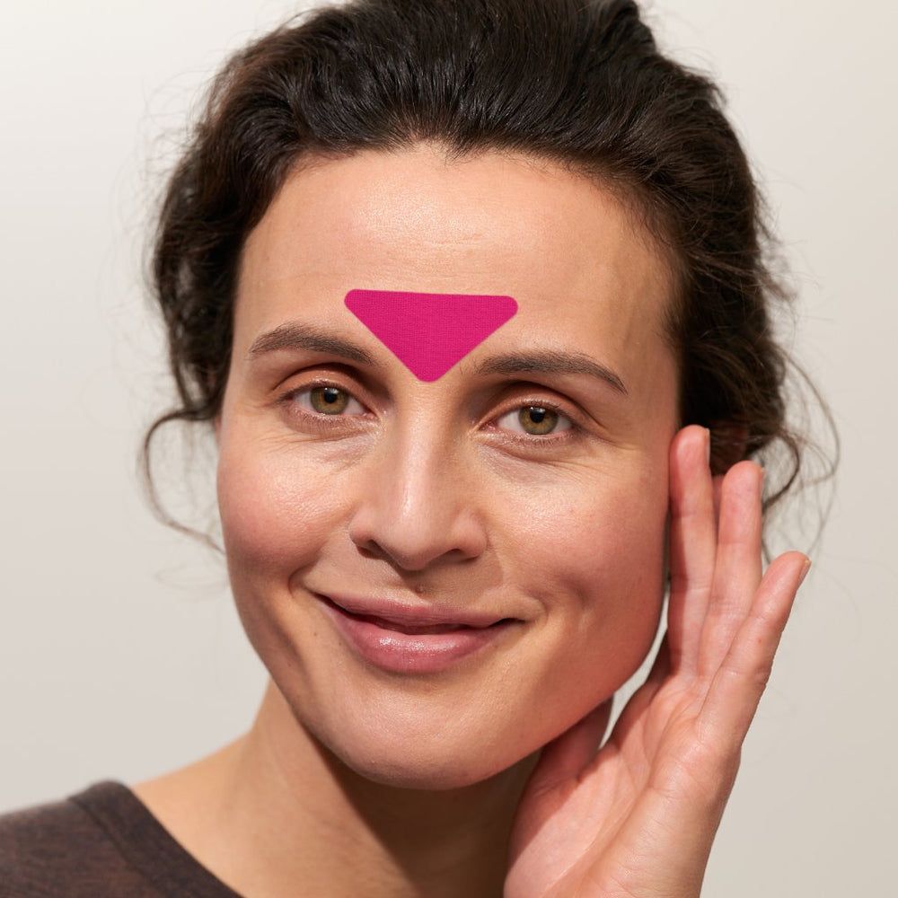 Gros plan sur une femme avec un patch facial rose sur le front. Le patch est triangulaire. La femme sourit.