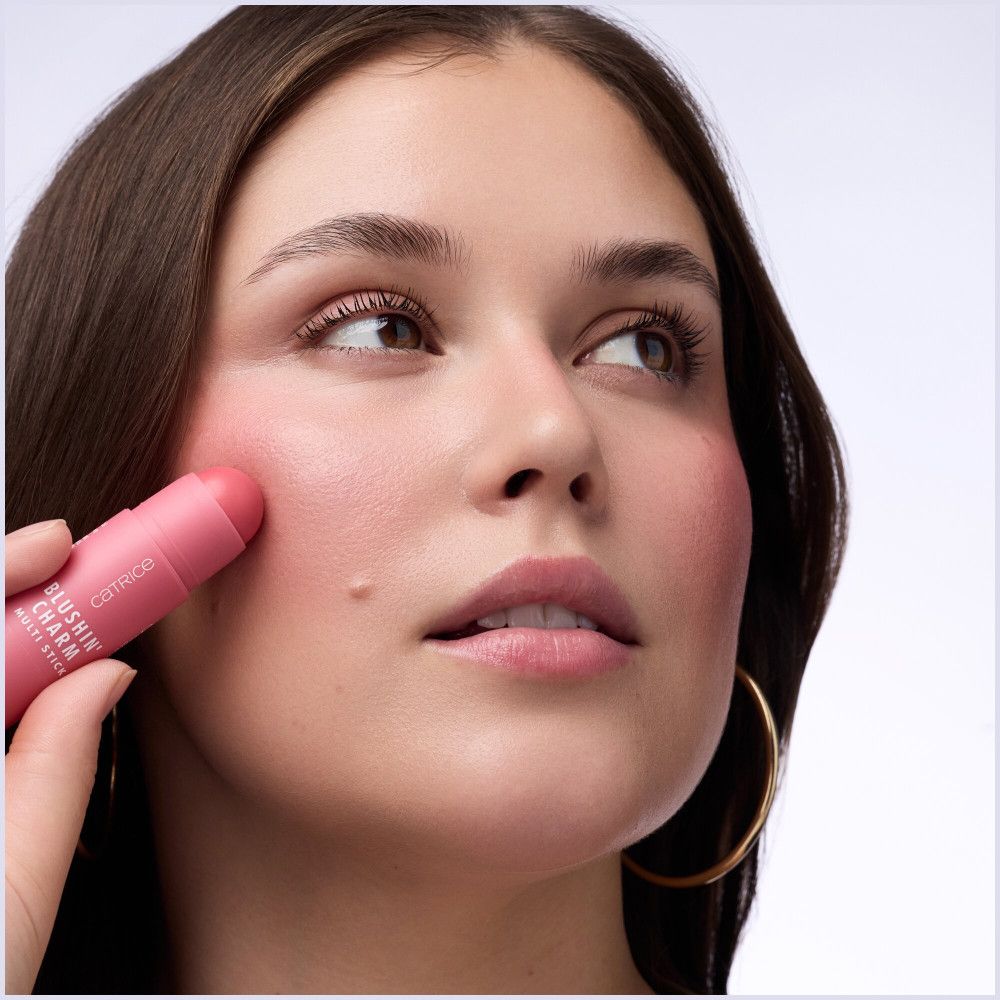 Femme appliquant Catrice Blushin' Charm Multi Stick sur la joue. Stick rose, inscription : Catrice, Sunshin' Charm, Multi Stick.