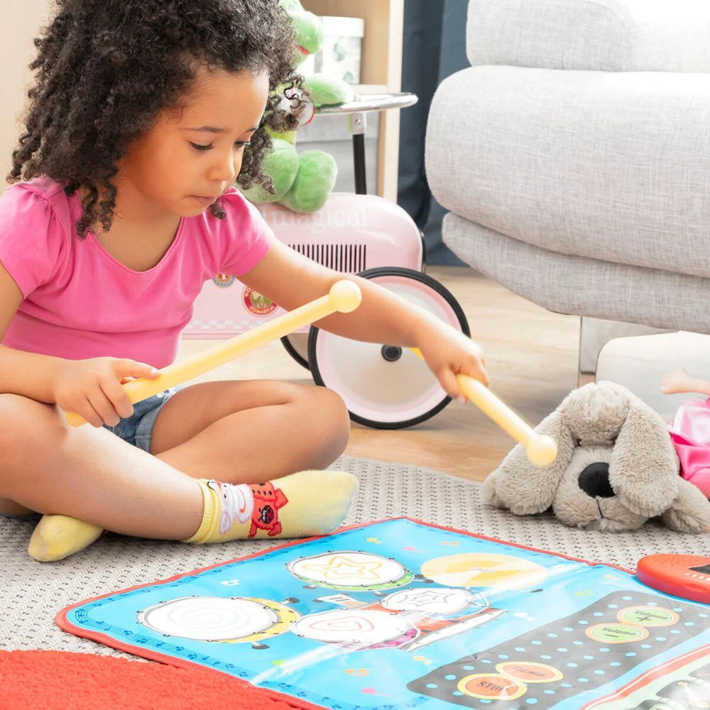 Un enfant frappe sur un tapis musical avec des baguettes. Un chien en peluche et des jouets sont en arrière-plan. Le tapis a des tambours.