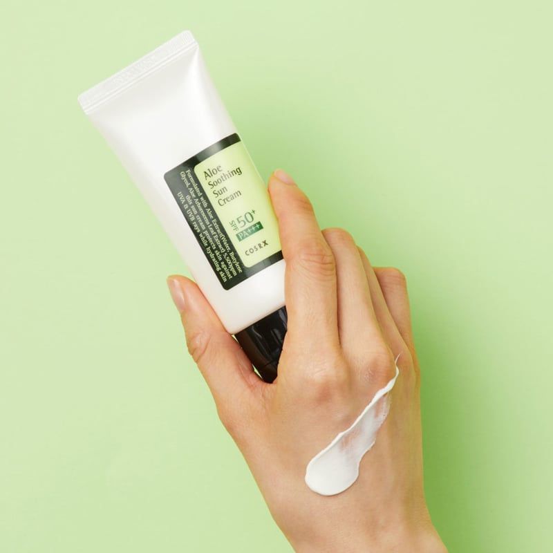 Hand houdt tube vast. Crème op hand. Tekst: Aloe Soothing Sun Cream SPF50+ PA+++. Zwarte dop.