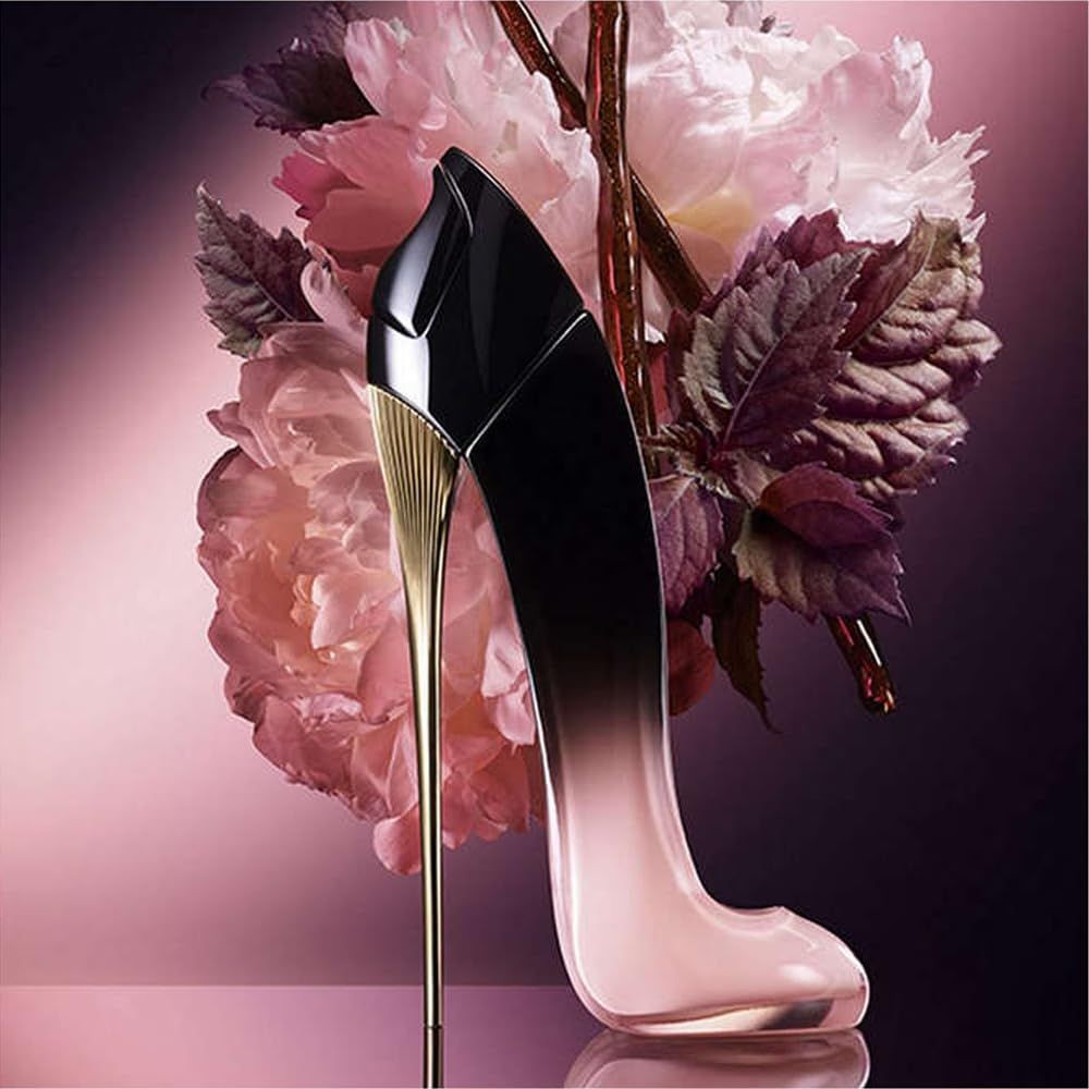 Flacon de parfum en forme de chaussure. Dégradé noir-rose, talon doré. Placé devant des fleurs roses.