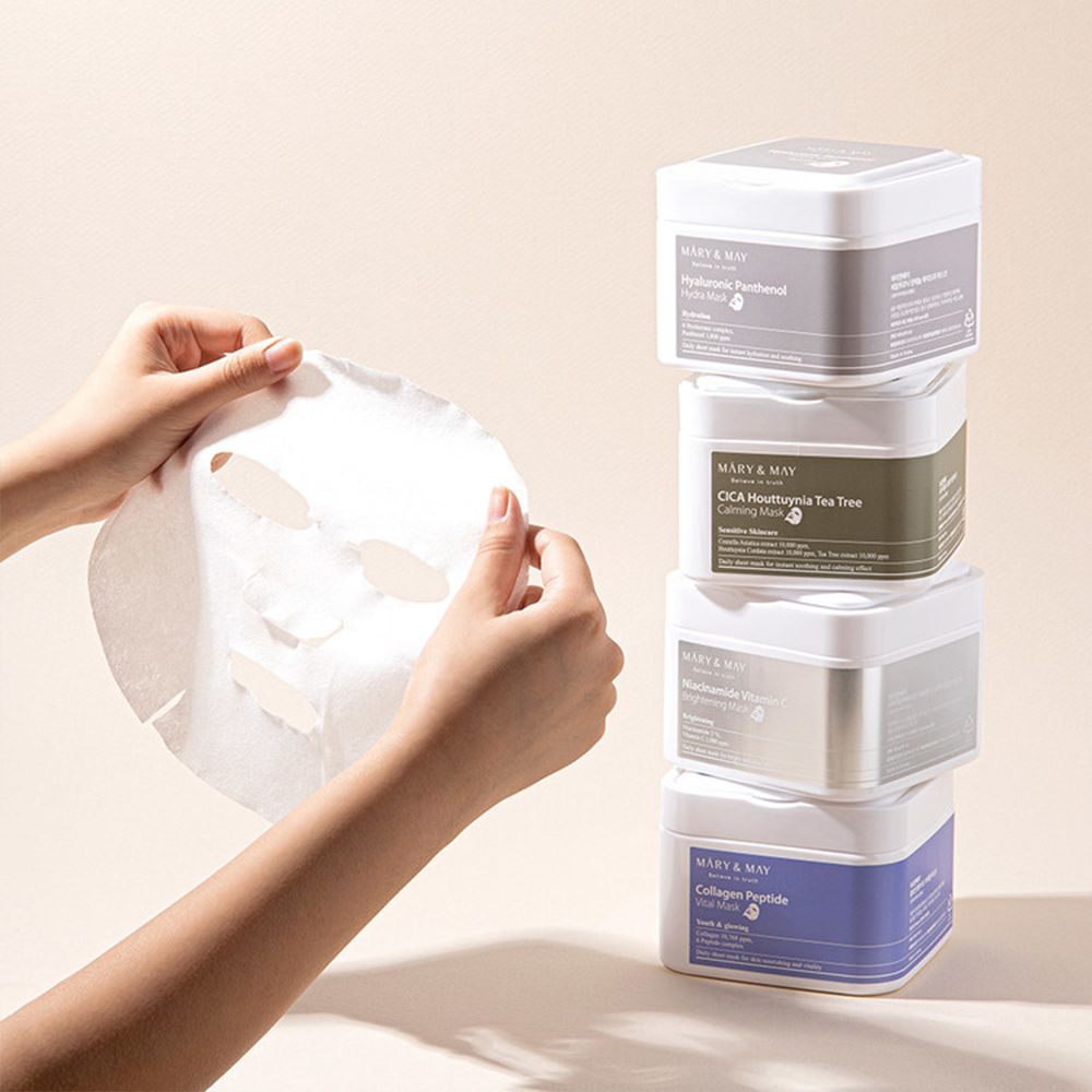 Handen houden een gezichtsmasker vast. Stapel MARY & MAY maskerverpakkingen: Hyaluronic Panthenol, CICA, Niacinamide, Collagen Peptide.