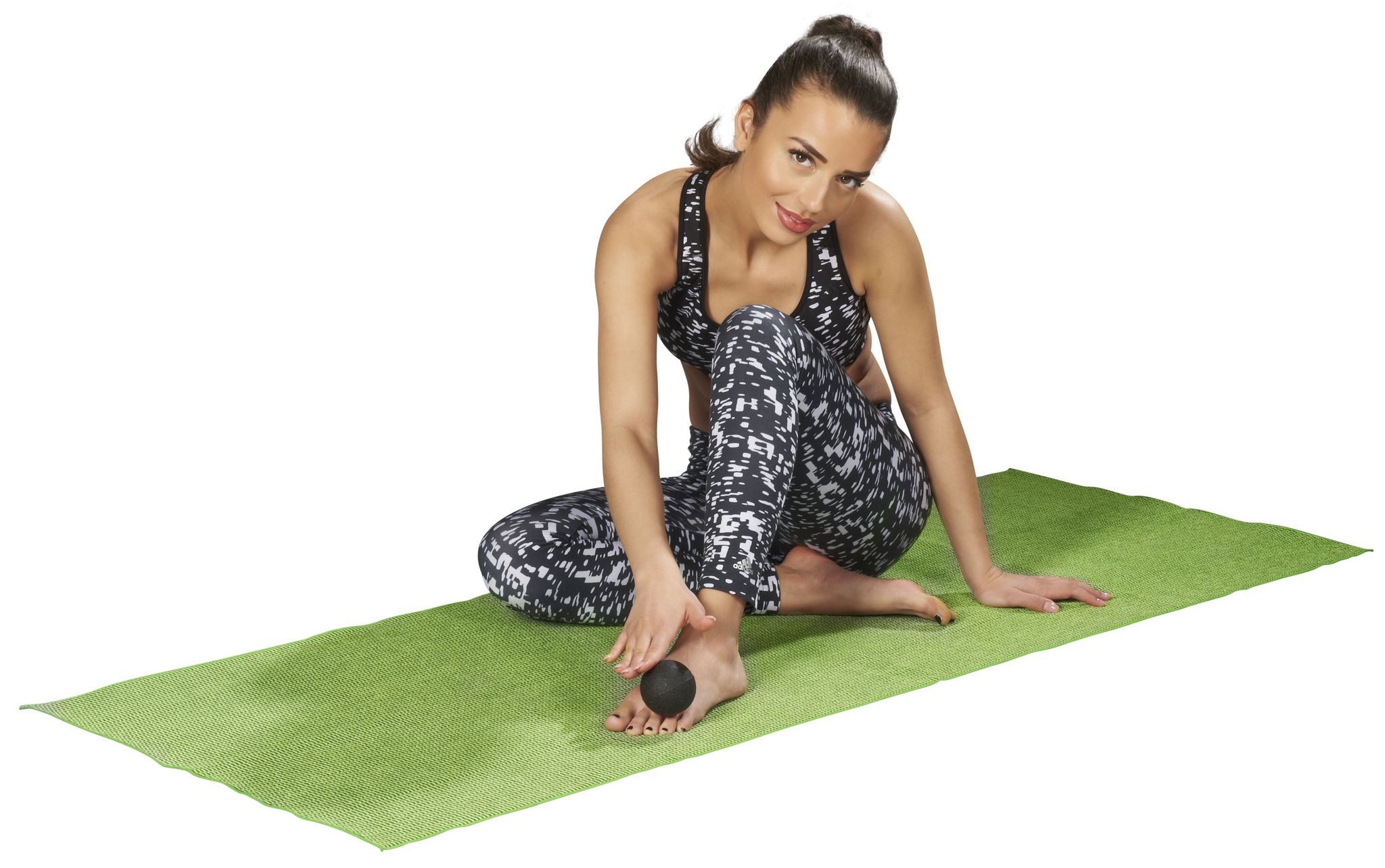 Femme assise sur un tapis vert. Balle noire sous le pied. Vêtements de sport : haut et pantalon à motifs. Regarde la caméra.
