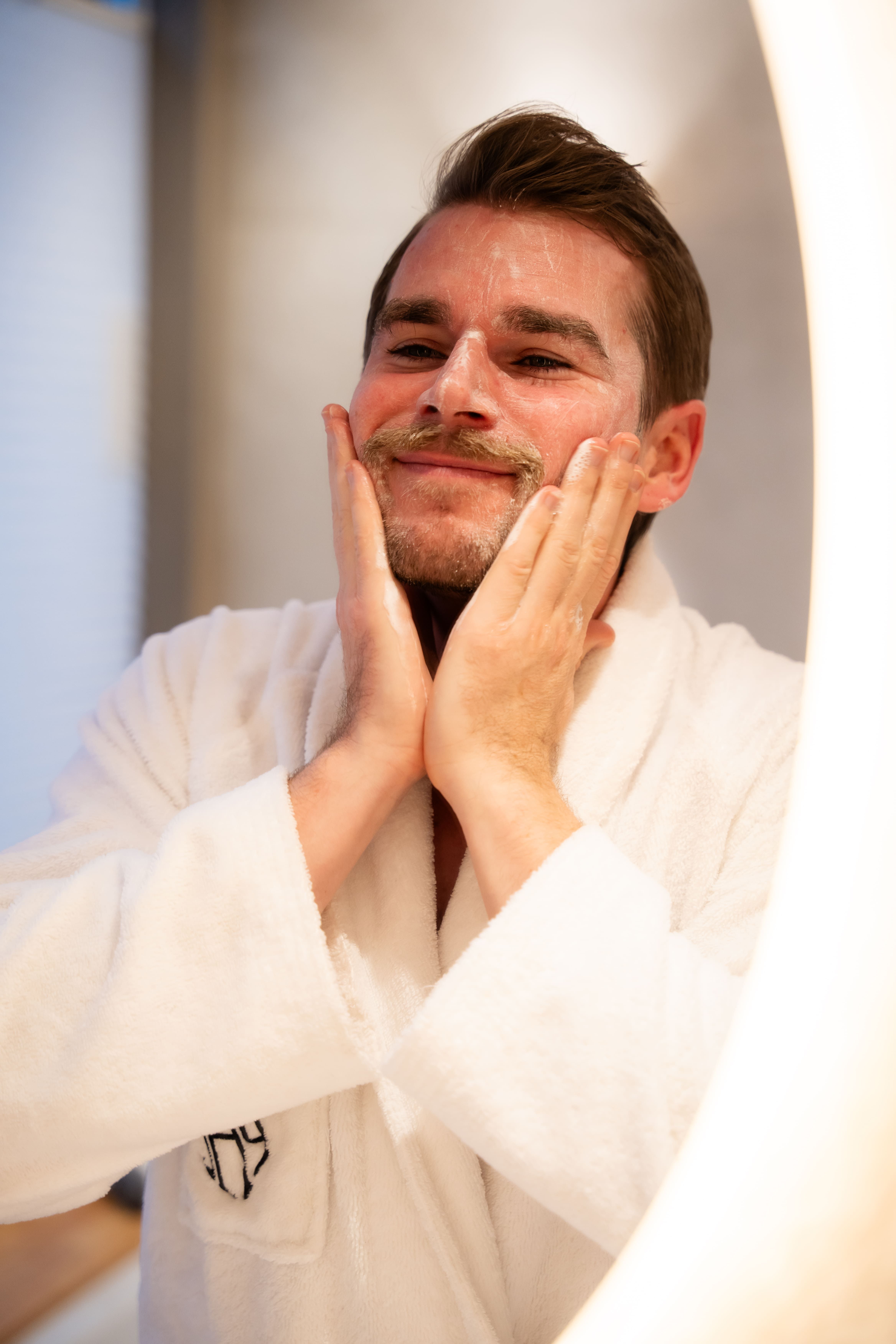 Man in witte badjas masseert gezicht voor verlichte spiegel.
