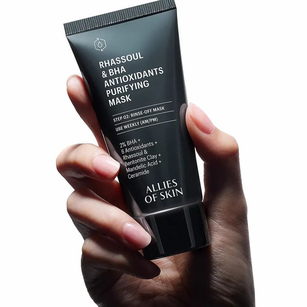 Donkergrijze tube in hand. Opschrift: Rhassoul & BHA Antioxidants Purifying Mask. Merk: ALLIES OF SKIN.