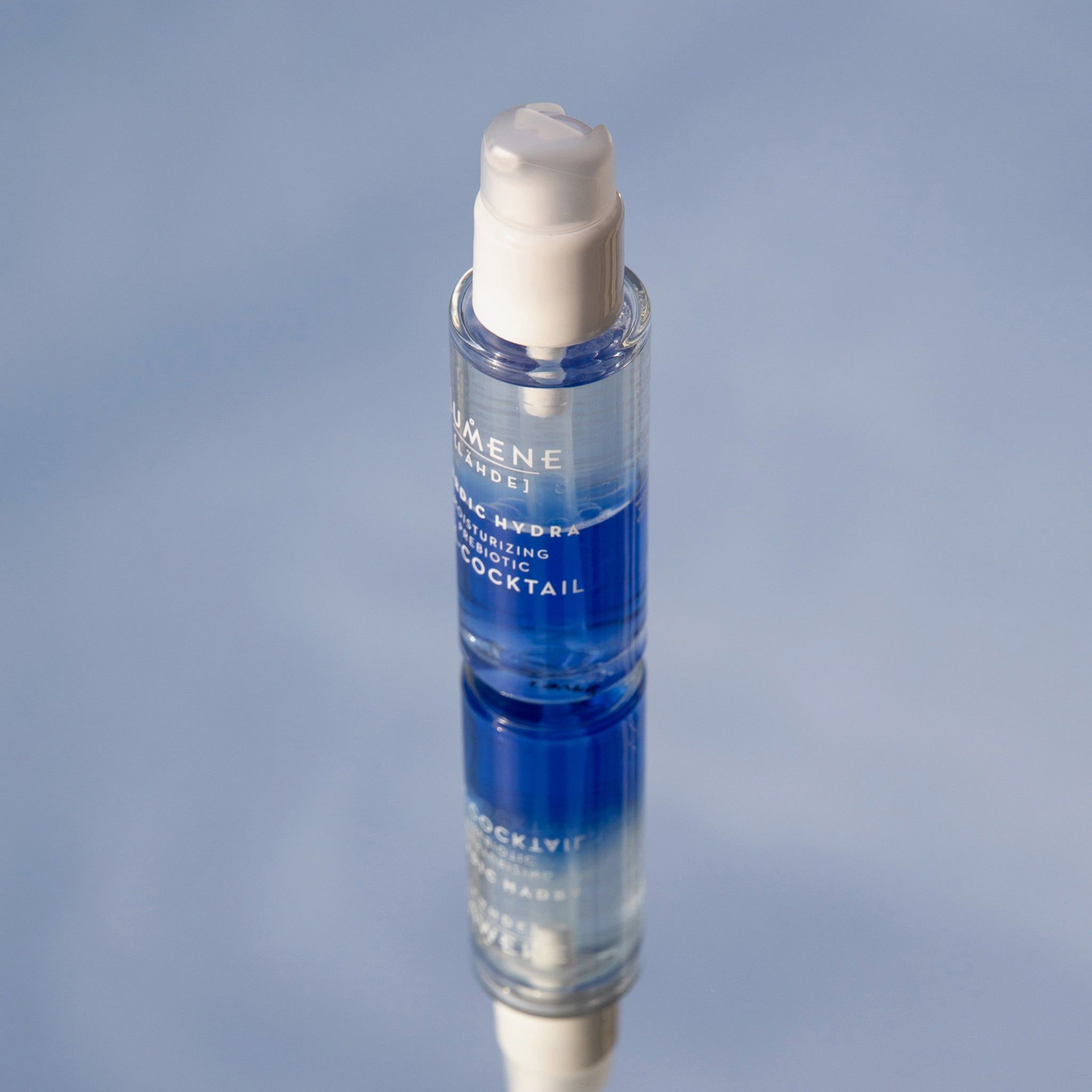 Flacon transparent avec pompe blanche, sur une surface réfléchissante. Liquide bleu et transparent visible. Nom du produit et marque.