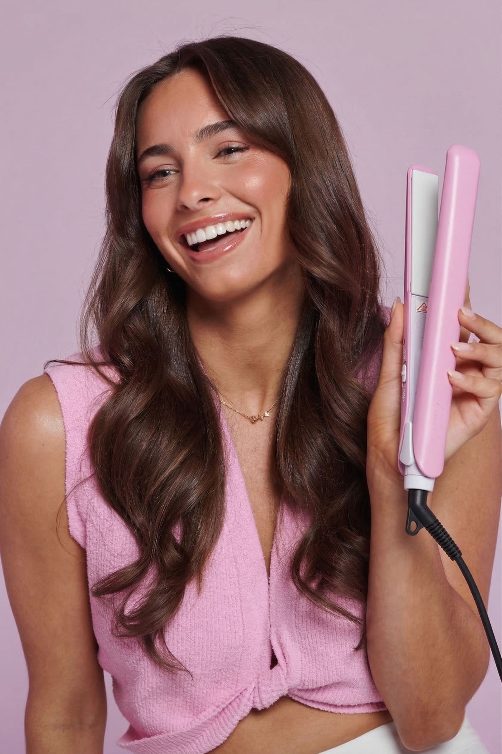 Vrouw houdt roze stijltang vast. Zwarte kabel. Vrouw lacht. Golvend bruin haar. Roze top.