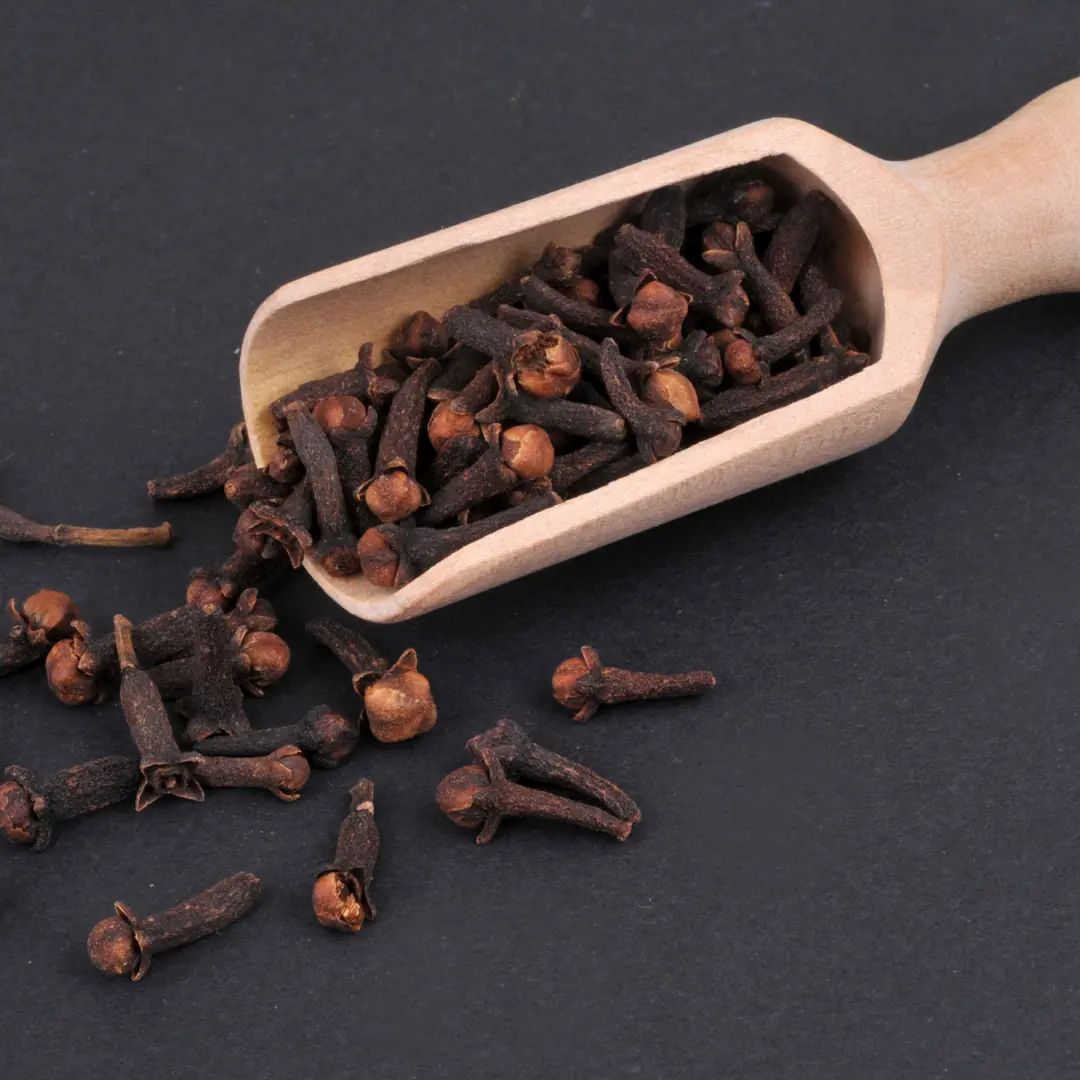 Kruidnagels in een houten schep op donkere ondergrond. Enkele kruidnagels verspreid.