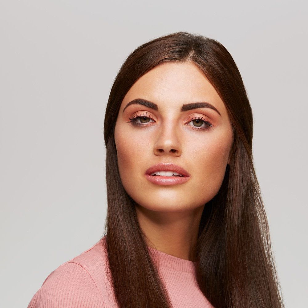 Portrait d'une femme aux longs cheveux bruns. Elle porte des vêtements roses et a les sourcils maquillés.