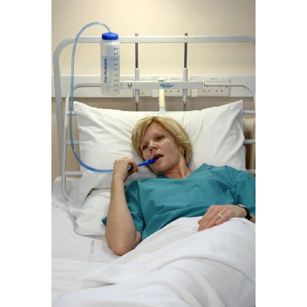 Vrouw in bed, drinkt uit een drinkfles met rietje. De fles is aan het hoofdeinde van het bed bevestigd.