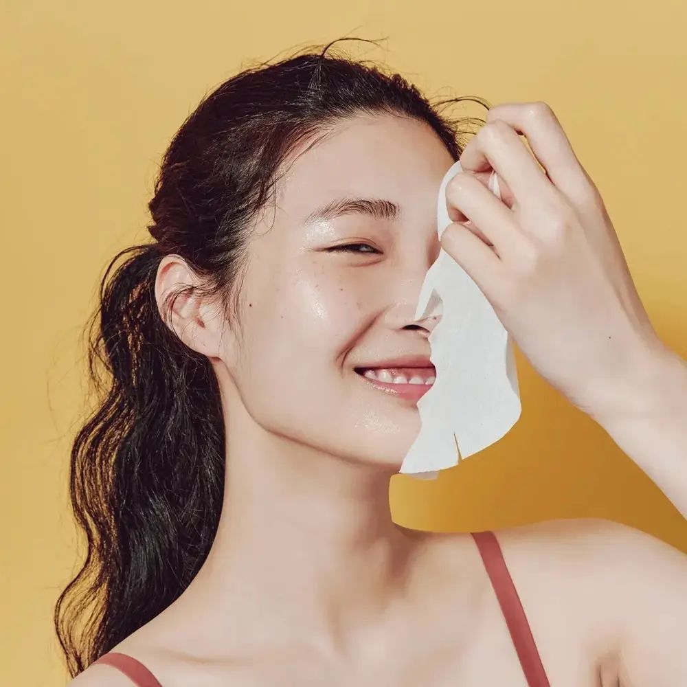 Persoon houdt gezichtsmasker voor gezicht. Masker bedekt helft van gezicht. Gele achtergrond. Persoon glimlacht.