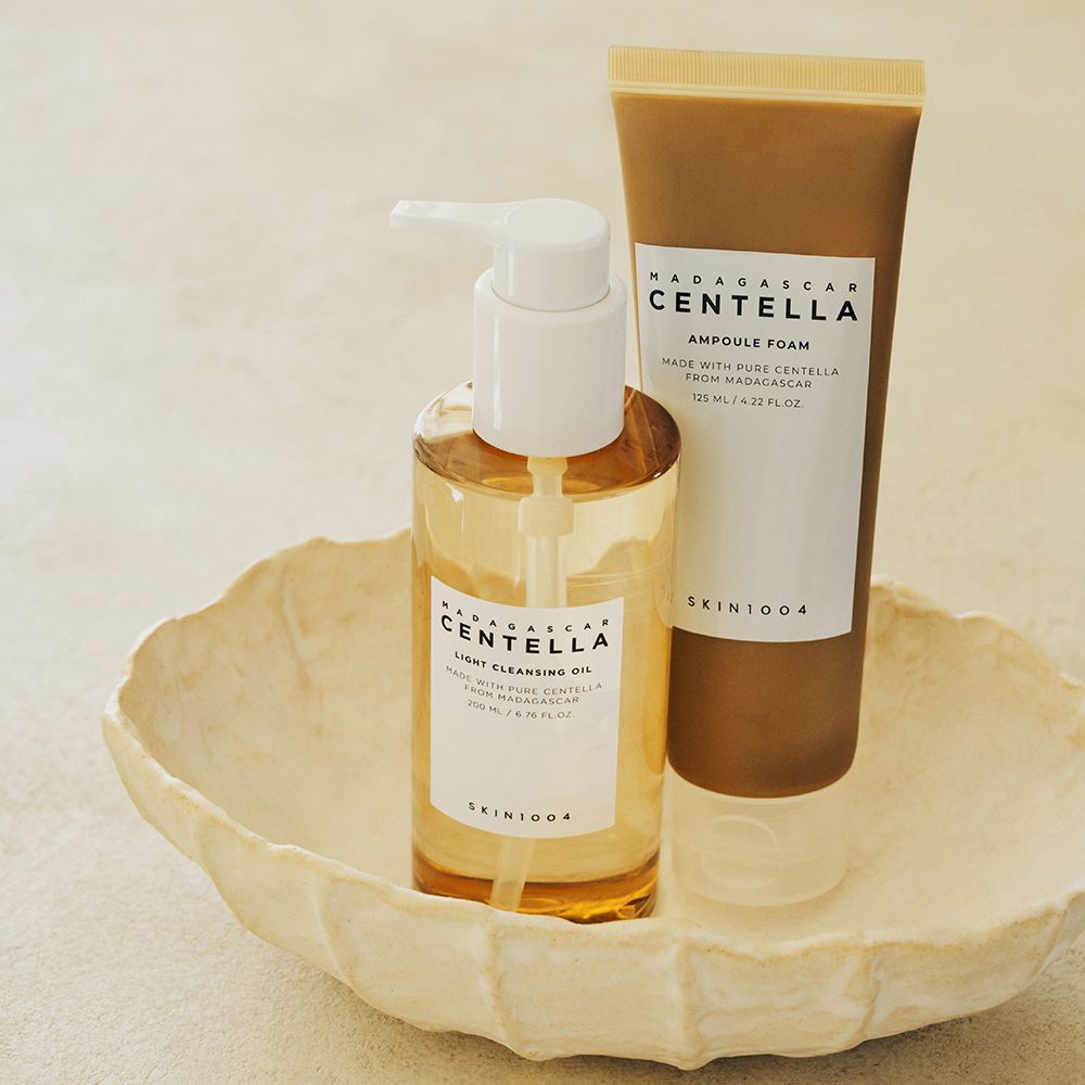 Deux produits sur un bol crème. À gauche, une bouteille d'huile nettoyante avec pompe, à droite, un tube de mousse ampoule. Les deux produits portent la marque SKIN1004.