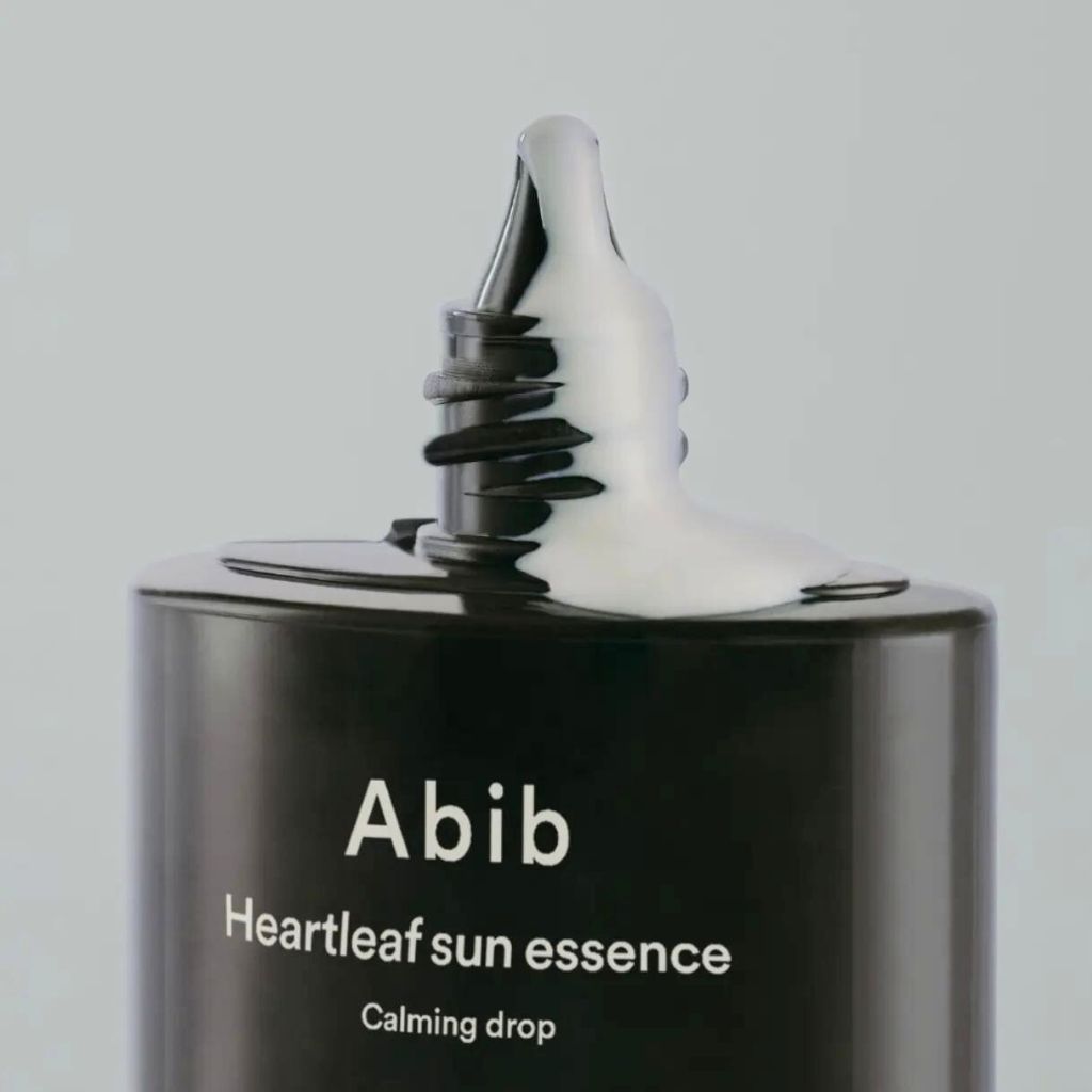 Close-up van een zwarte fles waar witte crème uitkomt. Opschrift: Abib Heartleaf Sun Essence Calming Drop.