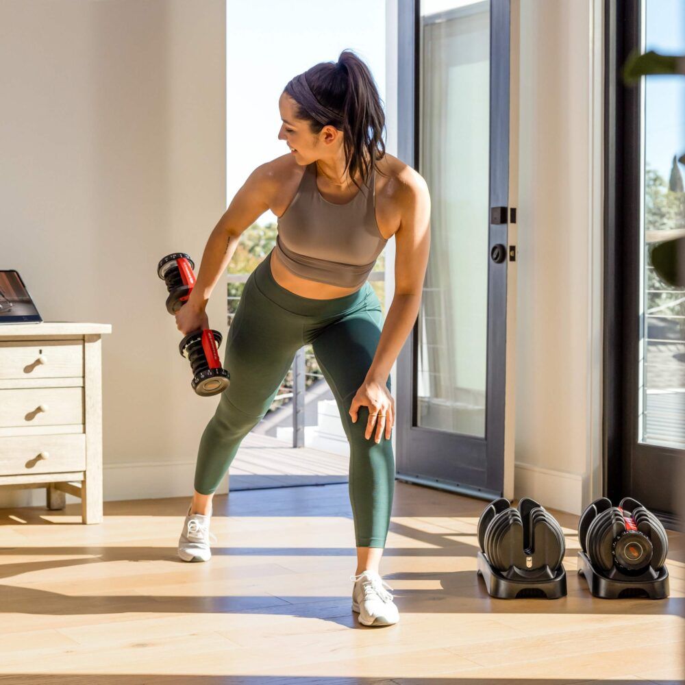Vrouw traint met een dumbbell. Twee andere dumbbells op de grond. Interieur met raam en meubels.