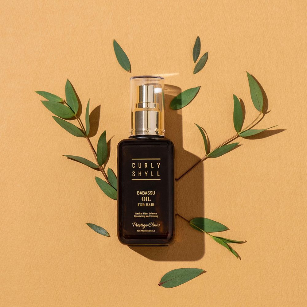 Bruine fles met gouden verstuiver. Opschrift: CURLY SHYLL, Babassu Oil for Hair. Omringd door bladeren.