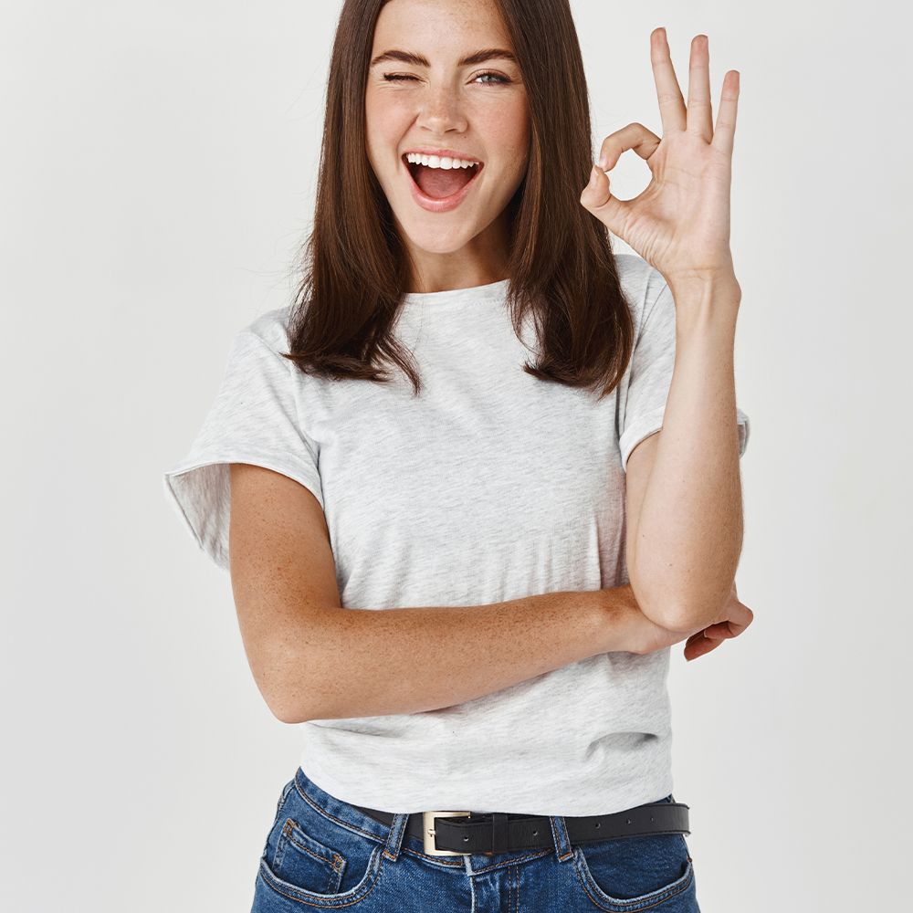 Une femme aux cheveux bruns et un t-shirt gris. Elle fait un clin d'œil et montre le signe 'OK' avec sa main.