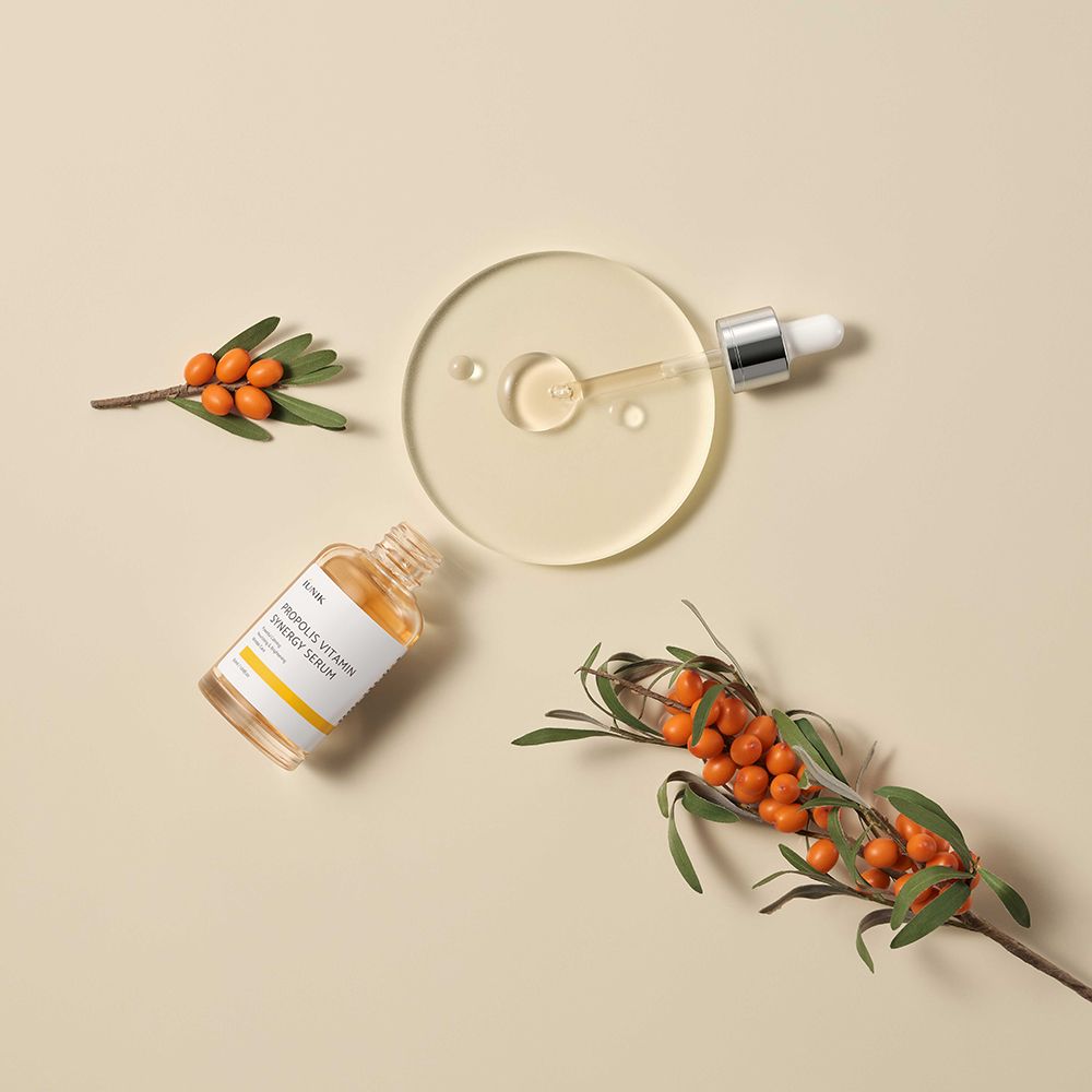 Serumfles met pipet, omringd door takken en bessen. Opschrift: iUNIK. Gele vloeistof zichtbaar.