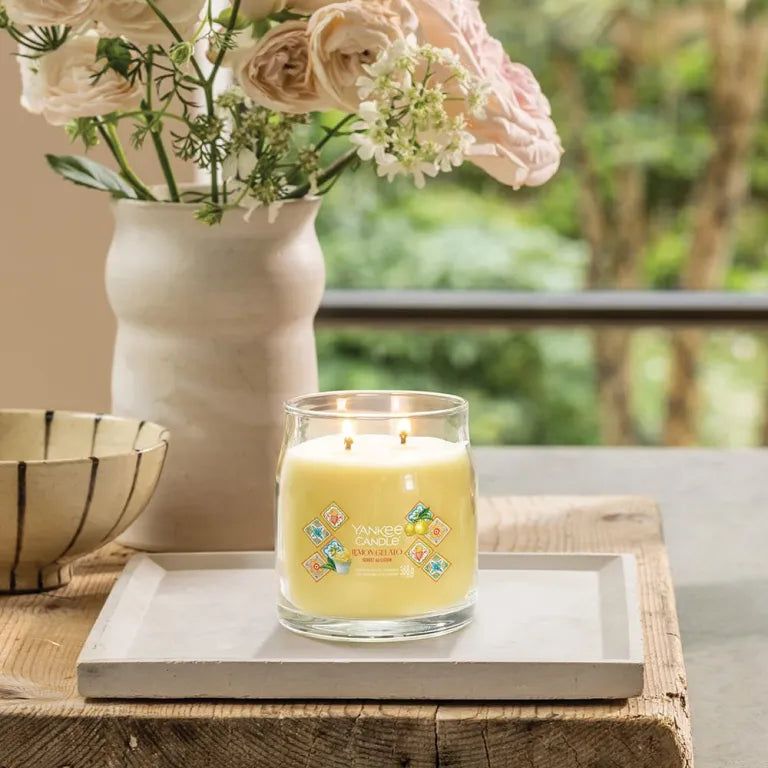 Bougie en verre avec cire jaune, deux mèches et étiquettes décoratives. Sur un plateau, entourée de fleurs et d'un vase.