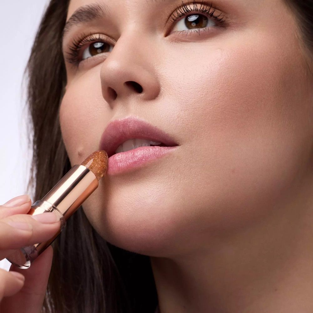 Femme appliquant du baume à lèvres. Baume à lèvres or rose. Lèvres légèrement ouvertes.