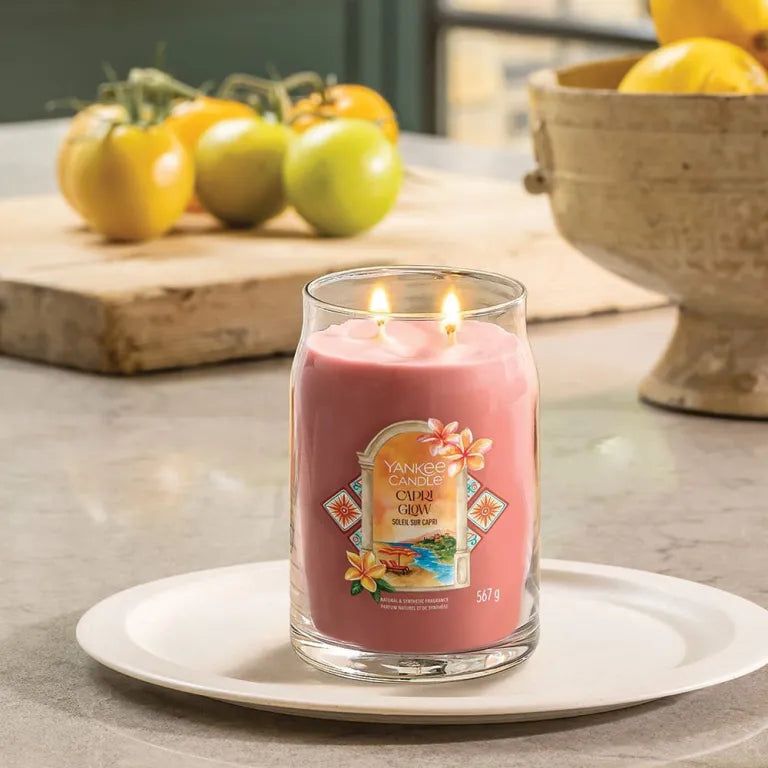 Bougie rose dans un verre, deux mèches, sur une assiette blanche. Inscription : Capri Glow. Fond : tomates.