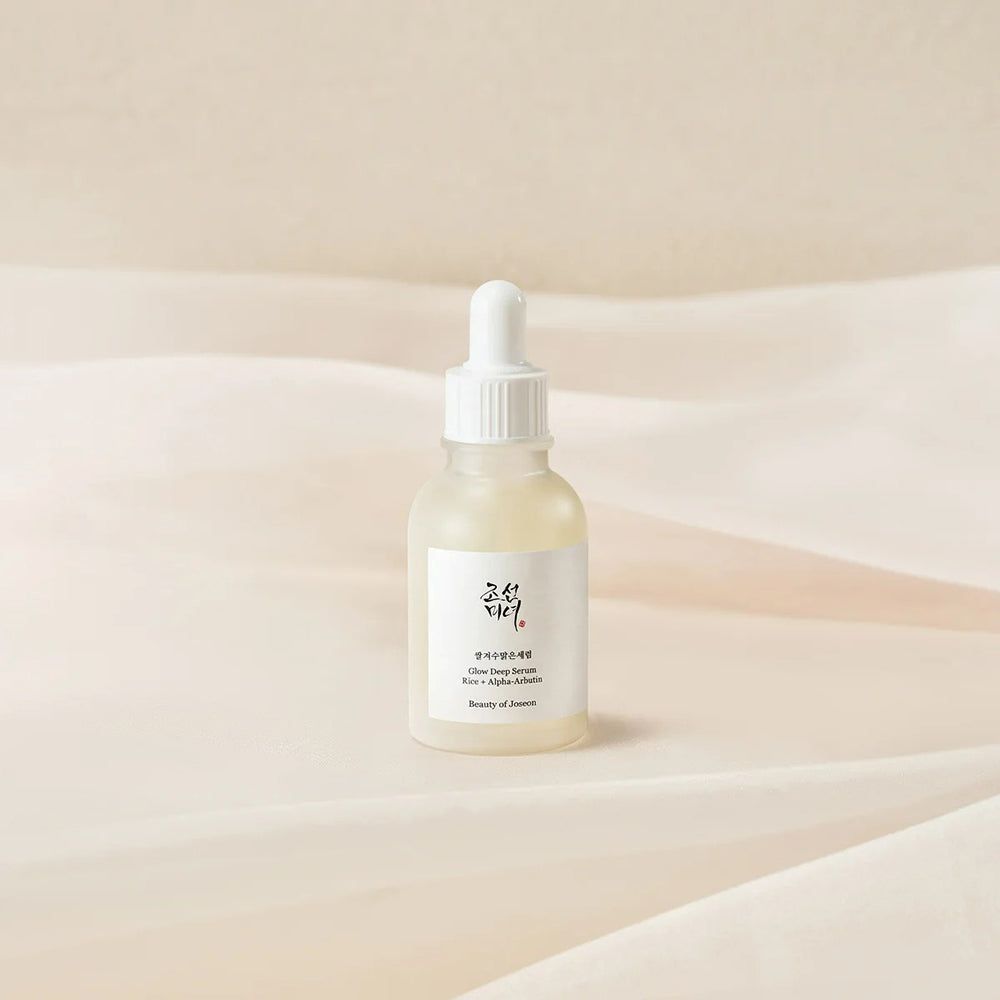 Serumfles met wit etiket. Opschrift: Glow Deep Serum, Rice + Alpha-Arbutin. Merk: Beauty of Joseon. Op lichte stof.